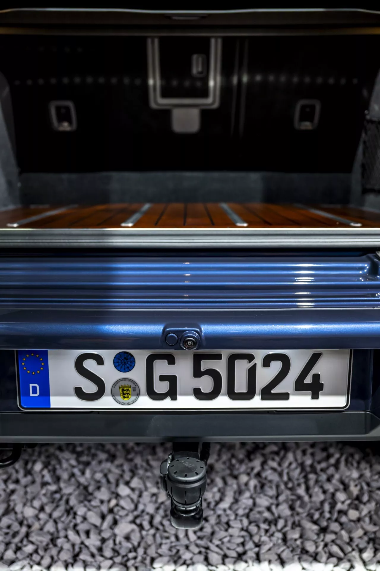 Szczegółowy zbliżenie na tylną część Mercedes-Benz G-Klasse 2024, prezentujące obszar bagażnika, luksusowe drewniane wykończenie bagażnika, wbudowaną kamerę cofania nad europejską tablicą rejestracyjną z niemieckim 'D' i hak holowniczy poniżej, wszystko uchwycone z niskiego, skierowanego do góry kąta, podkreślającego premium metaliczny lakier pojazdu i estetykę designu.
