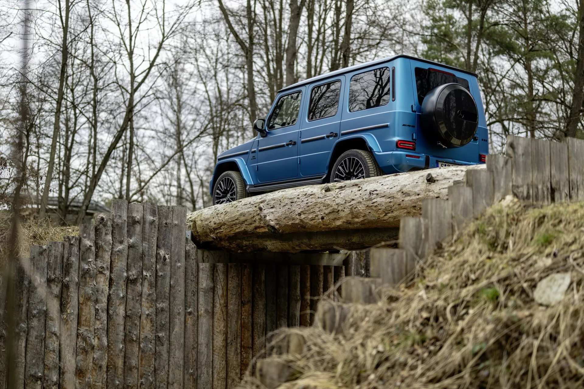 Mercedes-Benz klasy G 2024 w kolorze niebieskim, widok z boku podkreślający jego kwadratowy design, zawieszenie terenowe i prześwit na podwyższonej powierzchni, ze skupieniem na jego trwałych i luksusowych cechach na tle gołych drzew i drewnianego płotu