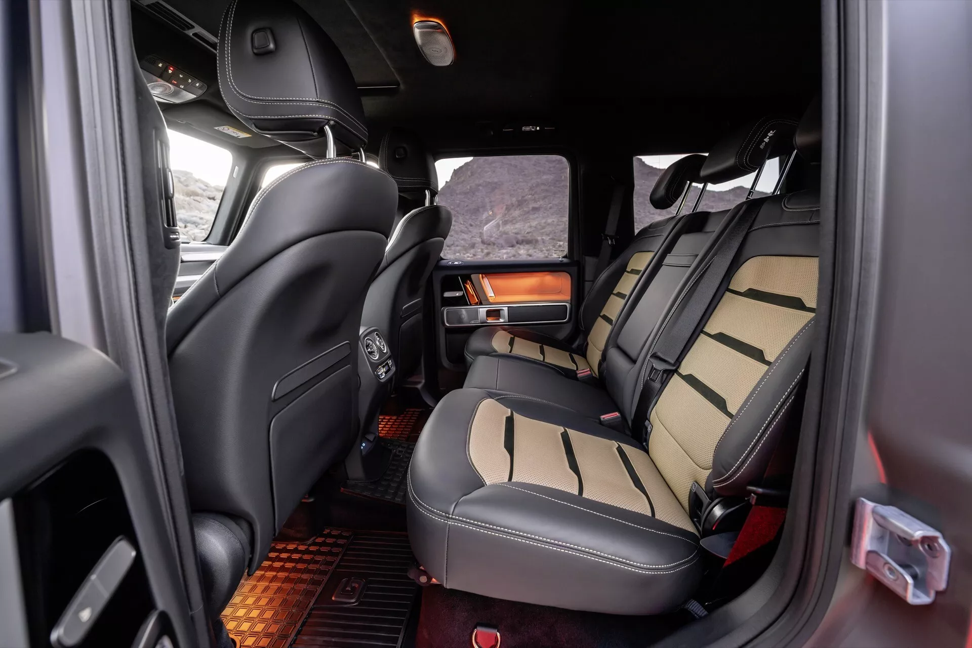 Wnętrze Mercedes-Benz Klasy G (G-Klasse) 2024 z widokiem na obszar siedzeń drugiego rzędu z siedzeniami tapicerowanymi dwukolorową skórą, składanym podłokietnikiem z uchwytem na napoje, widocznymi siedzeniami trzeciego rzędu i luksusowymi detalami, w tym elementy drewniane i oświetlenie ambientowe