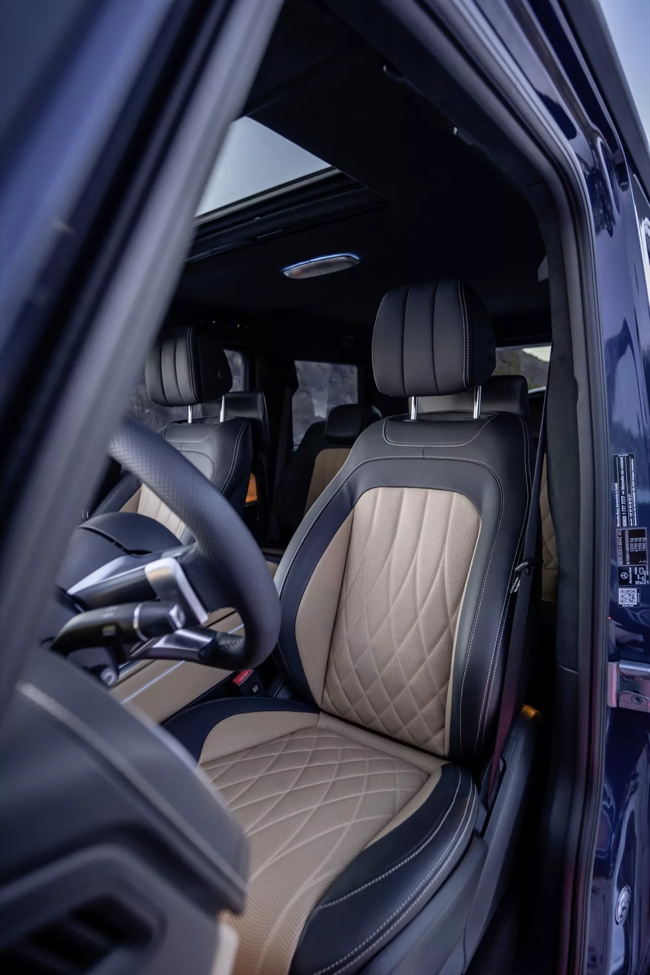 Widok wnętrza Mercedes-Benz Klasy G 465 2024 od strony kierowcy z otwartymi drzwiami, przedstawiający luksusową tapicerkę i materiały wysokiej jakości, ergonomiczną kierownicę i zaawansowane elementy designu