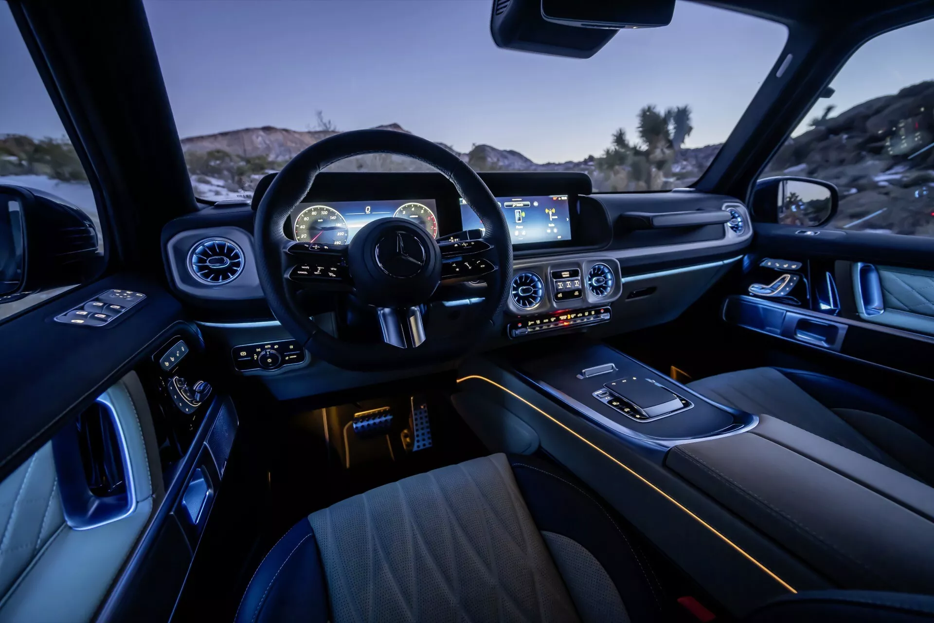 Widok wnętrza Mercedes-Benz G-Klasse 2024, podkreślający design kokpitu z perspektywy tylnej części pasażerskiej, prezentujący wielofunkcyjną sportową kierownicę, deska rozdzielcza z cyfrowym zestawem wskaźników, szerokoekranowy system infotainment, kontrolę klimatu, ambientowe oświetlenie wnętrza, luksusowe fotele przednie, selektor biegów, uchwyty na kubki, design drzwi z kontrolami elektrycznymi i materiały wykończeniowe premium.
