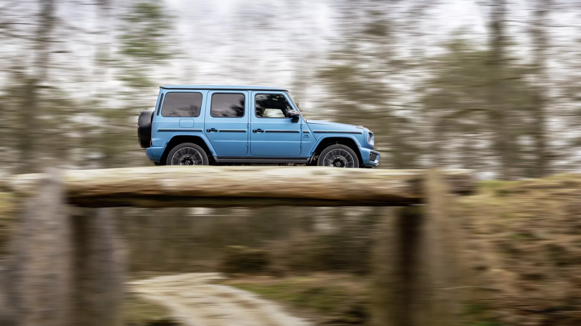 Poruszające się Mercedes-Benz G-Klasse (Klasa G) 465 z 2024 roku na drodze gruntowej, uchwycone techniką panningu, aby podkreślić ruch, pokazuje pełny widok boczny podkreślający terenowy design i felgi aluminiowe.