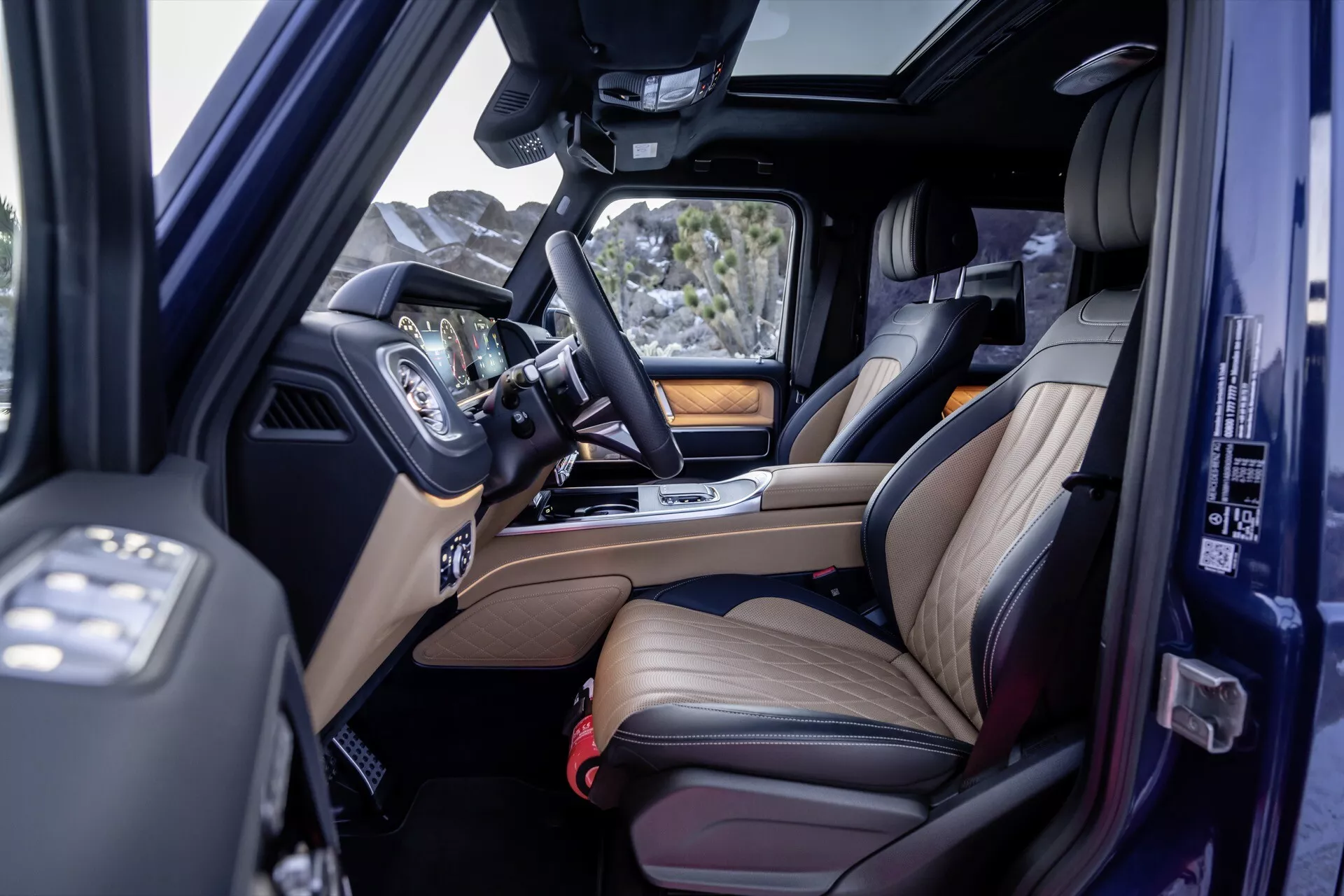 Widok wnętrza Mercedes-Benz Klasy G 2024 pokazujący przednie i tylne siedzenia skórzane z akcentami drewna, system infotainmentowy i dach panoramiczny z podniesionego kąta