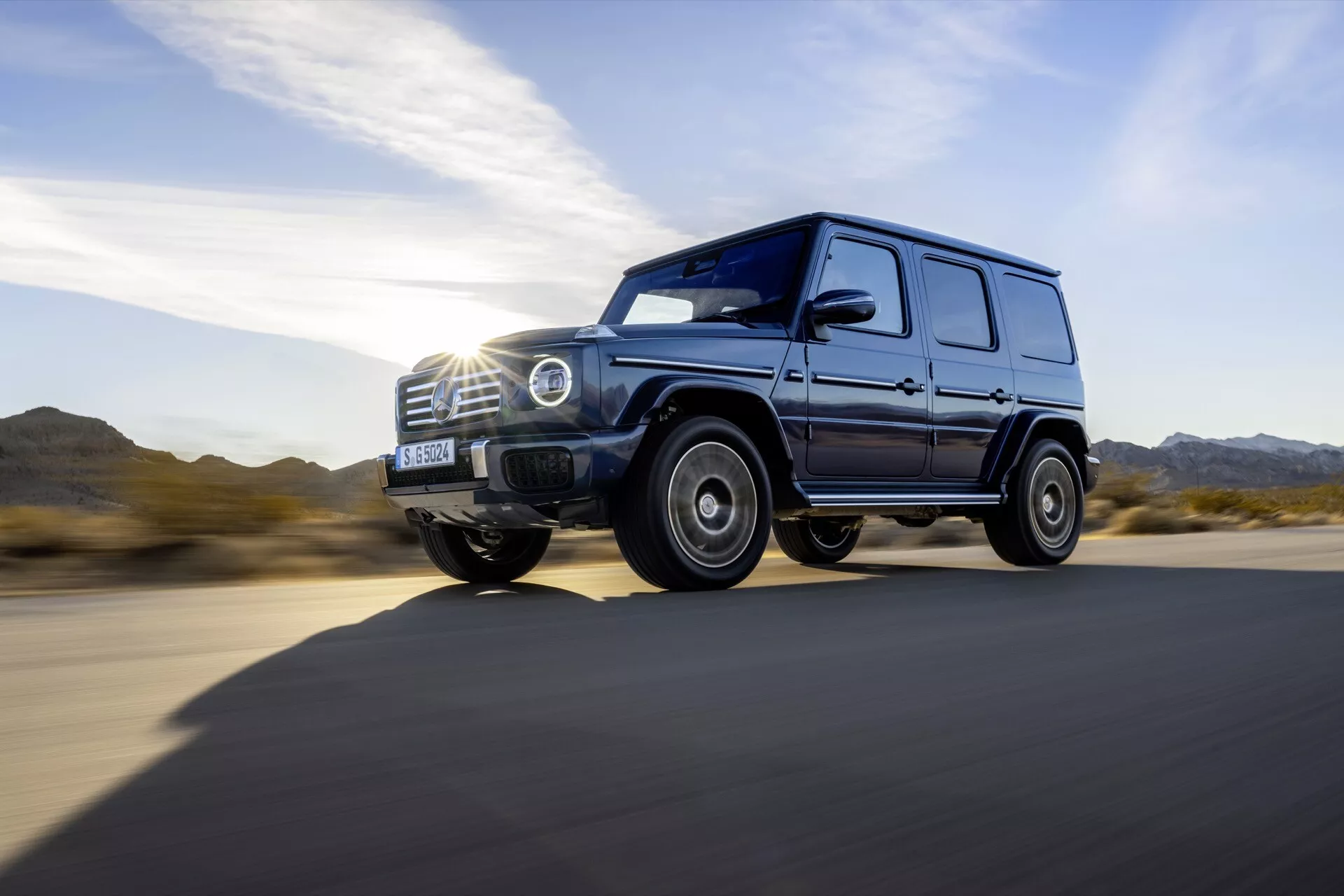 Luksusowy SUV Mercedes-Benz G-Class 465 z 2024 r. w dynamicznym widoku bocznym, jadący po asfaltowej drodze, z reflektorem LED, solidnym wzorem kół, wysokim prześwitem i lśniącym wykończeniem w świetle dziennym