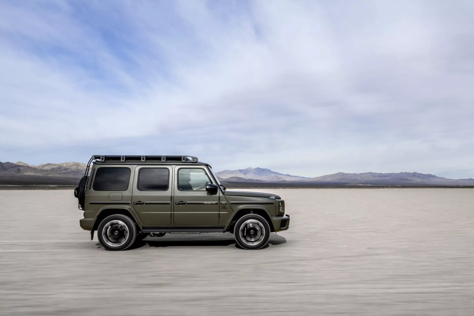 Mercedes-Benz G-Klasse 2024 w stylu wojskowego SUV-a, poruszający się po solniskach, pokazujący boczny i przedni profil z rozmyciem ruchu, podkreślający jego solidny design, zdolności terenowe i tradycyjne cechy bez widocznej nowoczesnej technologii