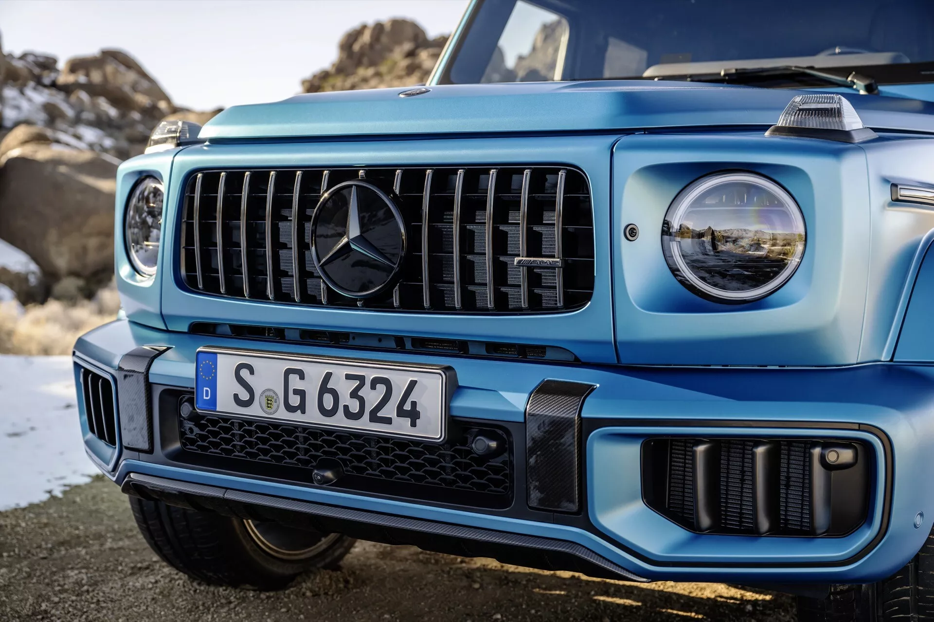 Zbliżenie na przednią lewą stronę jasnoniebieskiego Mercedes-Benz Klasy G z 2024 roku, przedstawiająca grill z pionowymi listwami i reflektory LED, ilustrujące zaawansowaną technologię oświetleniową i zdolności terenowe SUV-a