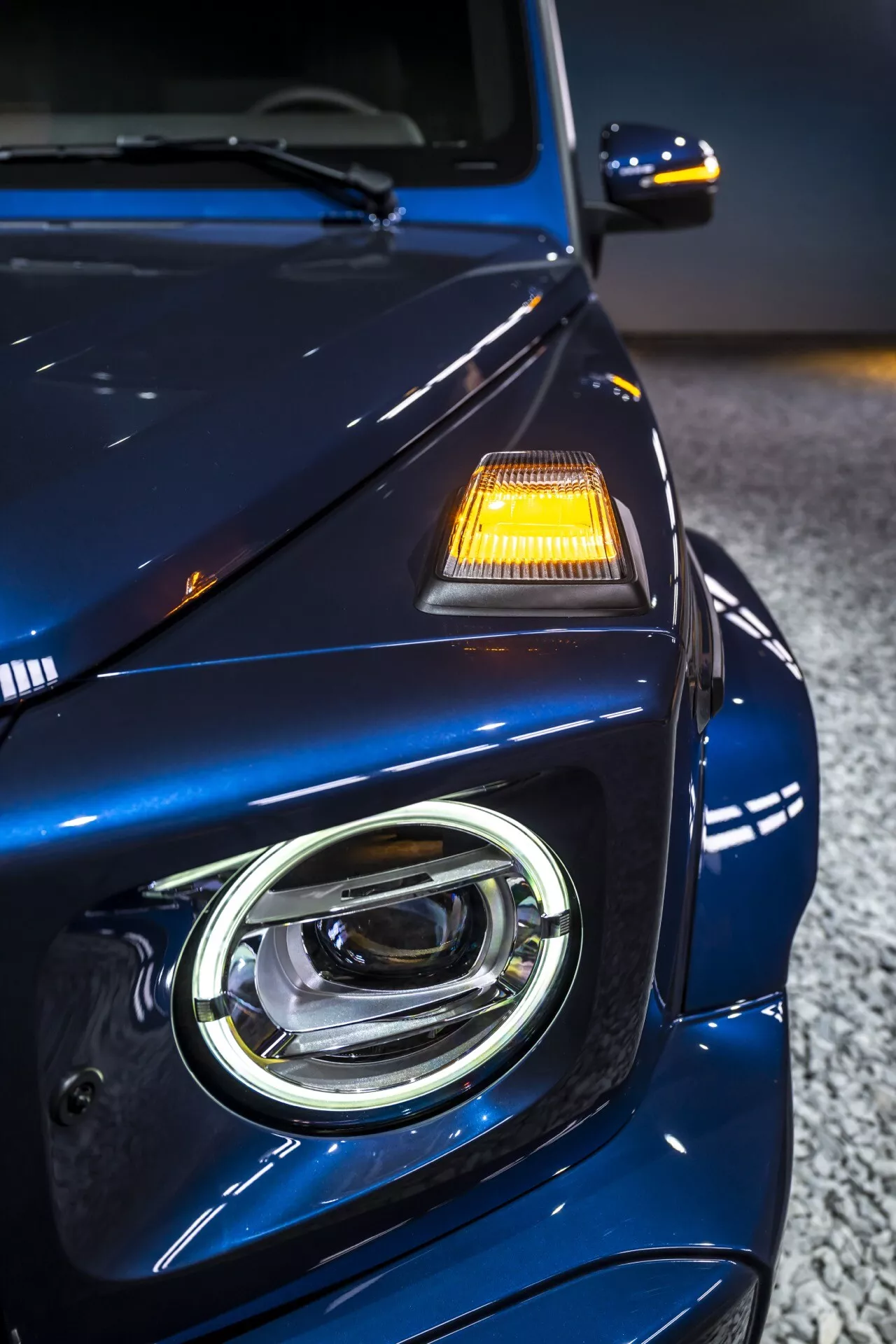 Zbliżenie na przedni lewy kwadrant Mercedes-Benz G-Klasse 2024, prezentujące projektowanie reflektorów LED, głębokie metaliczne niebieskie wykończenie premium i zaawansowane funkcje bezpieczeństwa