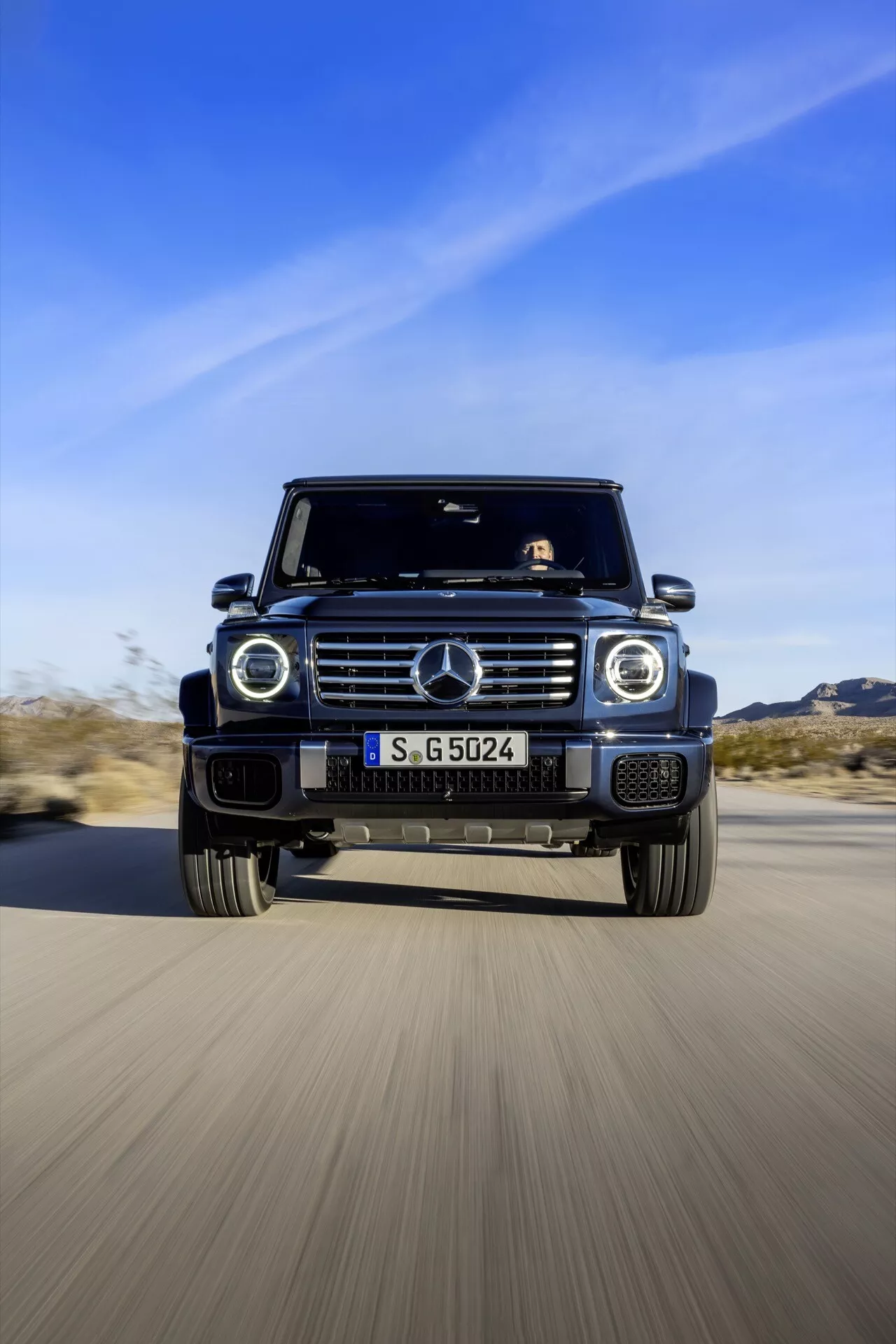 SUV Mercedes-Benz Klasy G 2024 w ruchu, ujęty z niskiego kąta czołowego, eksponujący jego agresywny design i zaawansowane funkcje jak charakterystyczna kratka, okrągłe reflektory, duże wloty powietrza i felgi ze stopów na tle pustyni