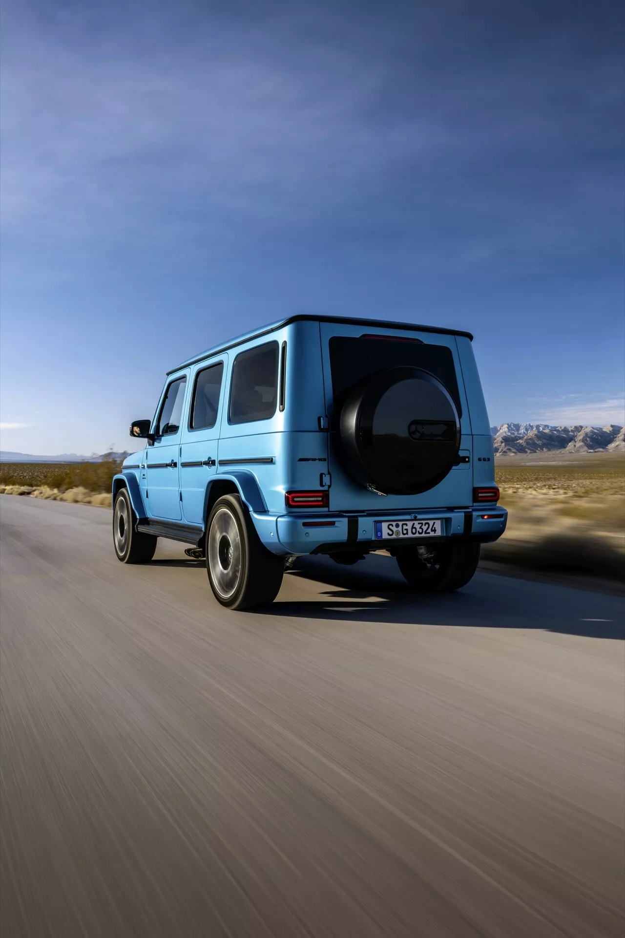 SUV Mercedes-Benz Klasy G 2024 w jasnym błękicie, uchwycony z tylnego, trzykwartalnego kąta pokazującego prawy profil, tylne koło, drzwi pasażera z tyłu, część dachu i zamontowaną oponę zapasową, na tle malowniczego krajobrazu z czystym niebem i górami, sugerujący przygodę i wydajność
