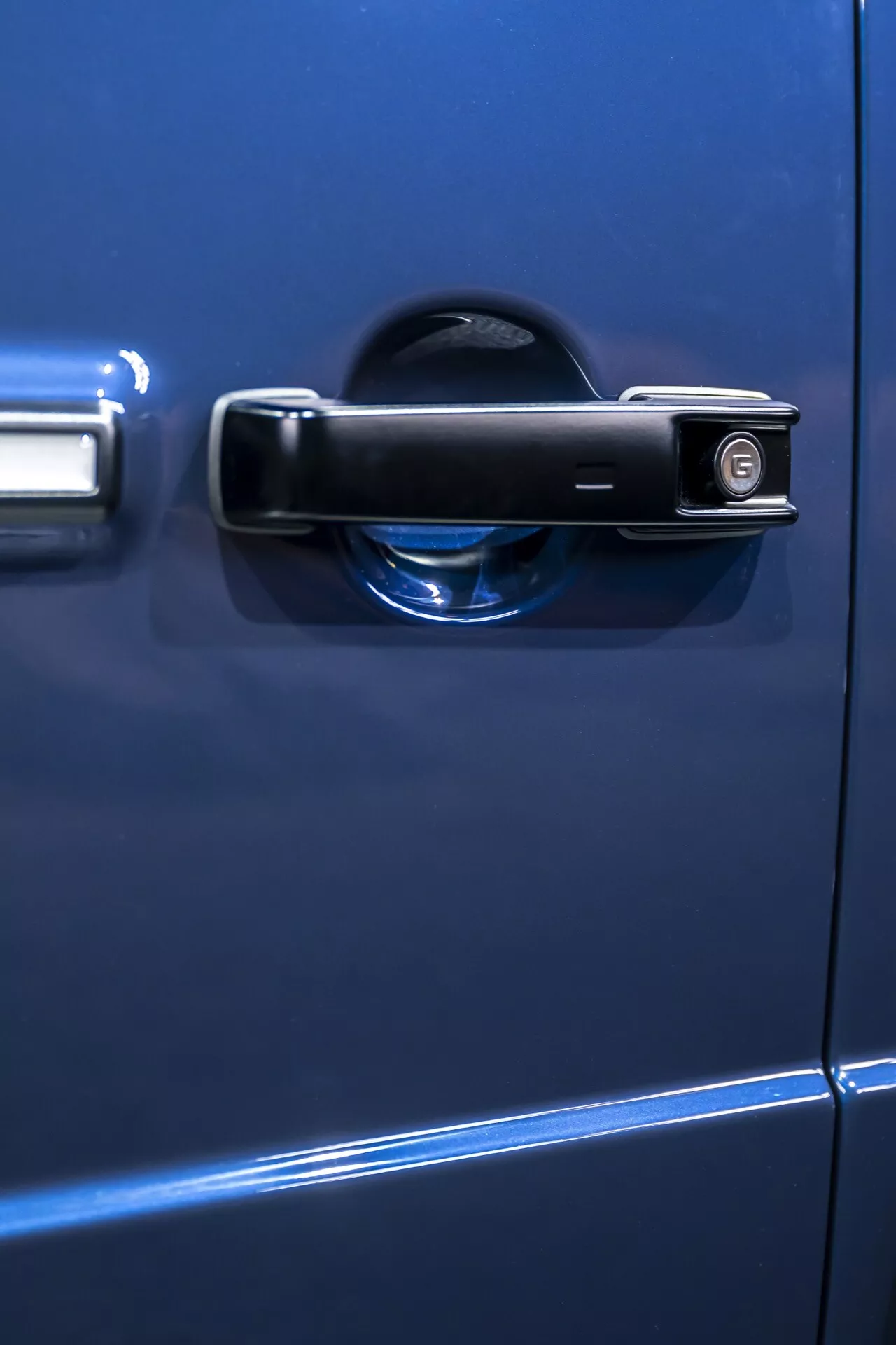 Zbliżenie na niebieską klamkę drzwi Mercedes-Benz G-Klasse 2024, prezentującą tradycyjny design z dziurką od klucza i wskaźnikiem zamknięcia, zdjęcie z boku o małej głębi ostrości