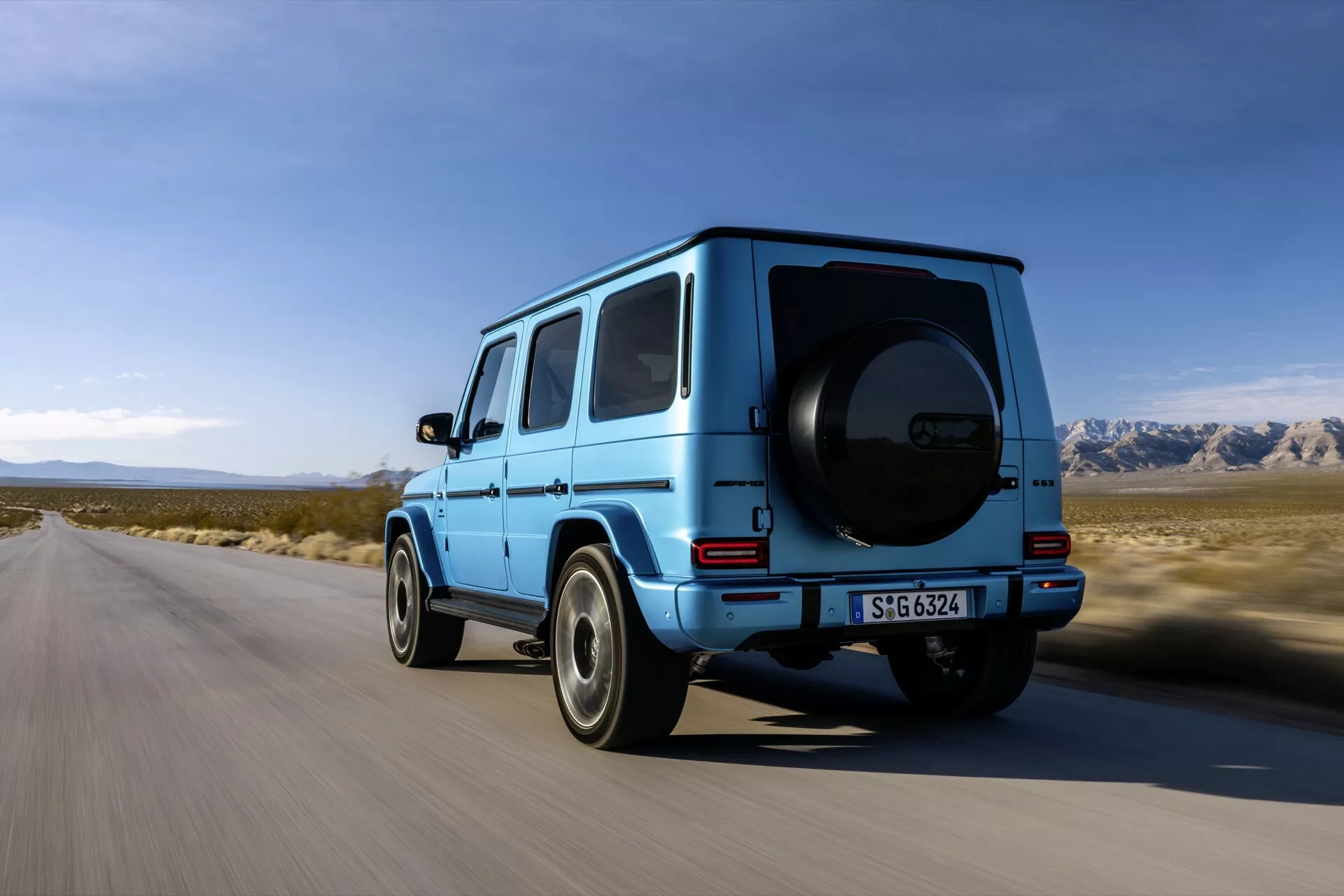 SUV Mercedes-Benz Klasa G rocznik 2024 w żywym niebieskim kolorze jedzie po utwardzonej drodze z półpustynnym górzystym krajobrazem i czystym niebem w tle, zdjęcie z niskiego do średniego poziomu terenu skupiające się na tylnej i częściowo bocznej stronie kierowcy pojazdu, charakterystyczny dla pojazdów terenowych i luksusowych pojazdów użytkowych kształt pudła, widoczna opona zapasowa montowana na tylnej klapie pojazdu, wyraźne nadkola i wysokie prześwit, koła z wieloma ramionami i widoczne zaciski hamulcowe, przyciemniane szyby z tyłu sugerujące ochronę UV i większą prywatność, tylne światła LED, i zderzak tylny utrzymany w kolorze karoserii