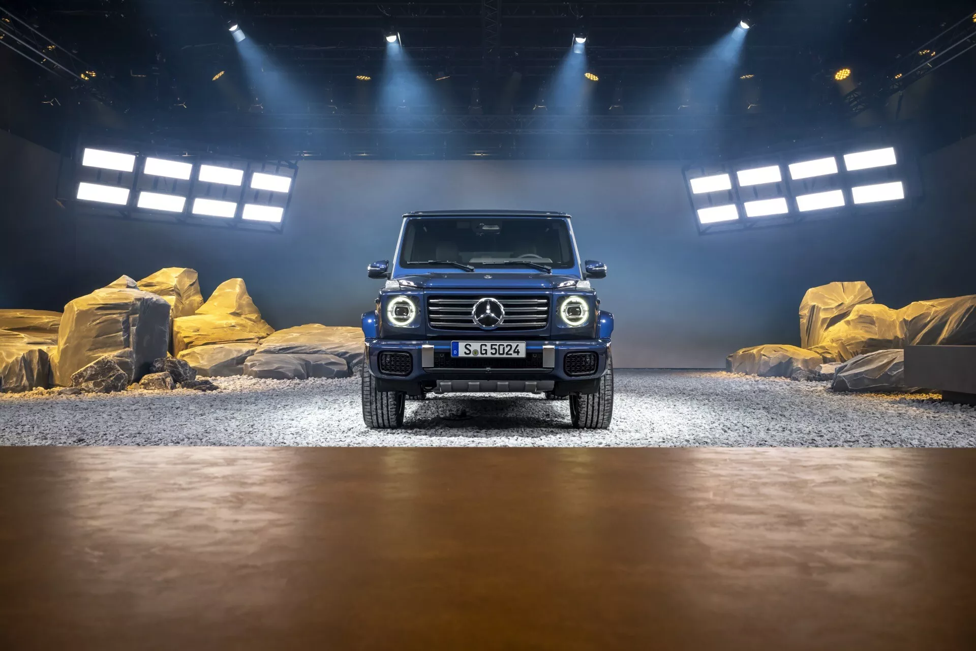 Mercedes-Benz Klasy G 2024 prezentowany w symulowanym wnętrzu off-road, widok z przodu podkreślający grill, reflektory LED i projekt zderzaka przedniego, z efektami oświetlenia studyjnego i naturalnego podkreślającymi jego metaliczny lakier i zdolności terenowe