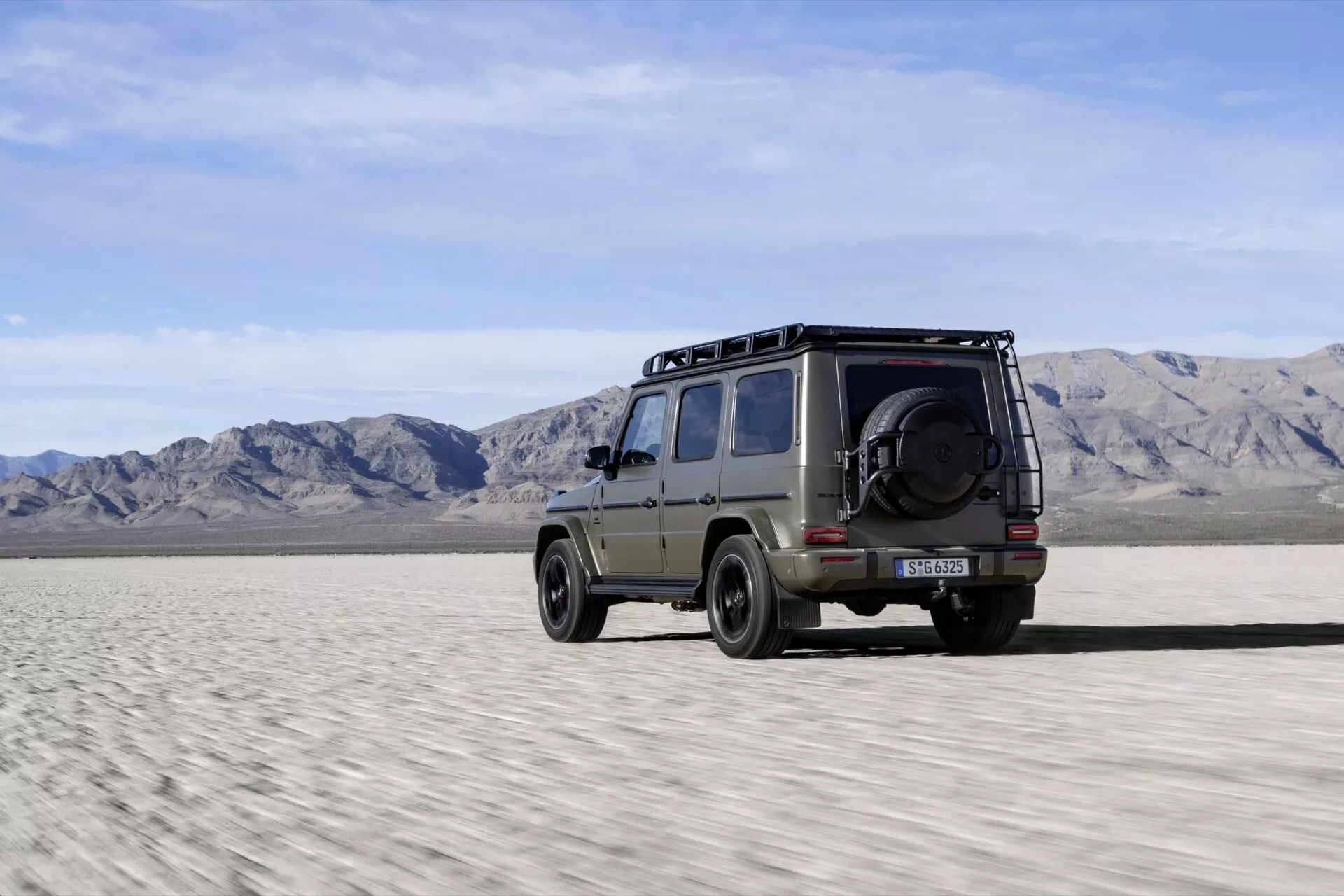 SUV Mercedes-Benz G-Klasse 2024 ukazujący swój profil boczny z nutką tyłu, prezentujący zdolności terenowe na pustynnym terenie, z relingami dachowymi, solidnymi oponami i kołem zapasowym zamontowanym na tylnych drzwiach