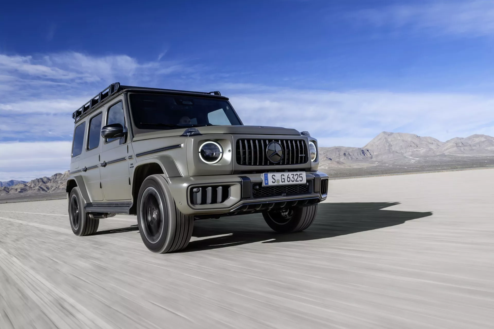 Srebrne SUV Mercedes-Benz Klasy G 2024, demonstrujące zdolności terenowe na płaskim, pustynnym terenie, z dynamicznym ujęciem z niskiego kąta podkreślającym jego robustną konstrukcję, okrągłe reflektory, przedni grill i ślad pyłu.
