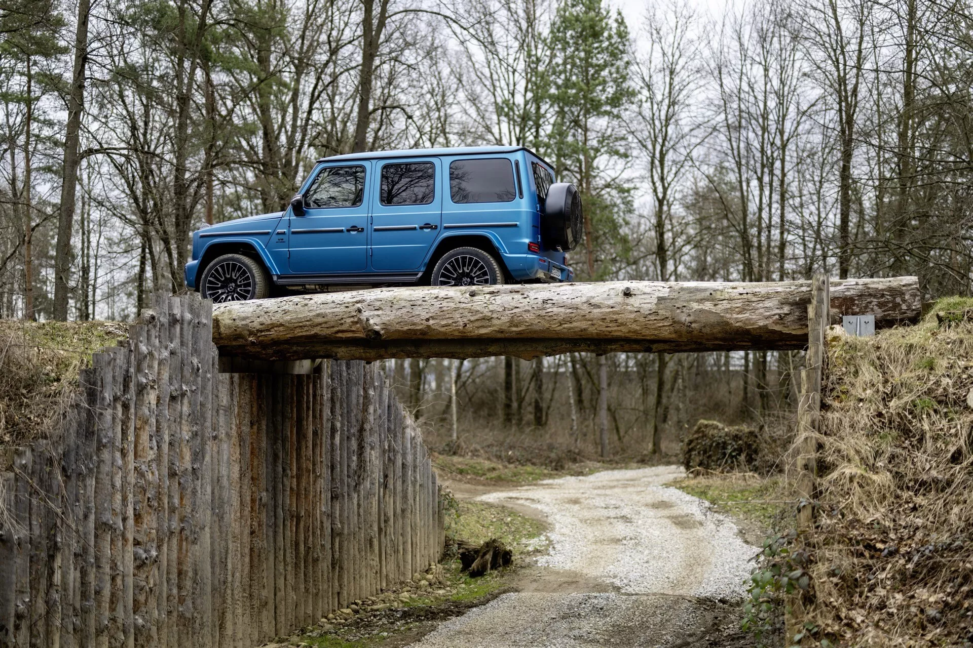 Mercedes-Benz Klasa G 2024 w jasnym niebieskim kolorze zaparkowany prostopadle na drewnianym belce w wiejskim otoczeniu, podkreślając jego kwadratowy sylwetka, wysokie prześwit, trwałe opony i cechy konstrukcyjne skoncentrowane na użytkowości, takie jak stopnie boczne, szyberdach i relingi dachowe na tle pochmurnego lasu