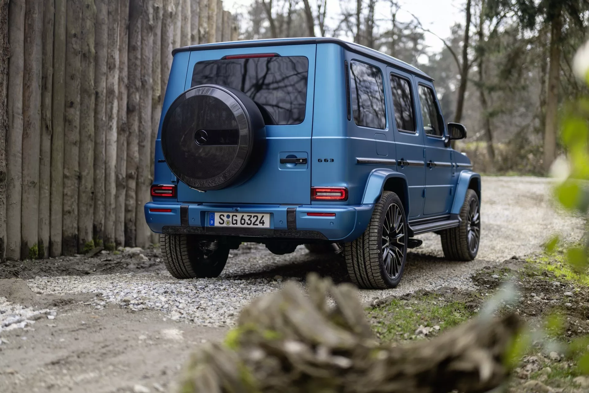 Tylny widok niebieskiego SUV-a Mercedes-Benz klasy G (G 465) 2024, podkreślający jego solidną, kanciastą konstrukcję, zamontowane koło zapasowe, tylne światła LED, wysoki prześwit i felgi z lekkich stopów na gruzowej ścieżce otoczonej naturą