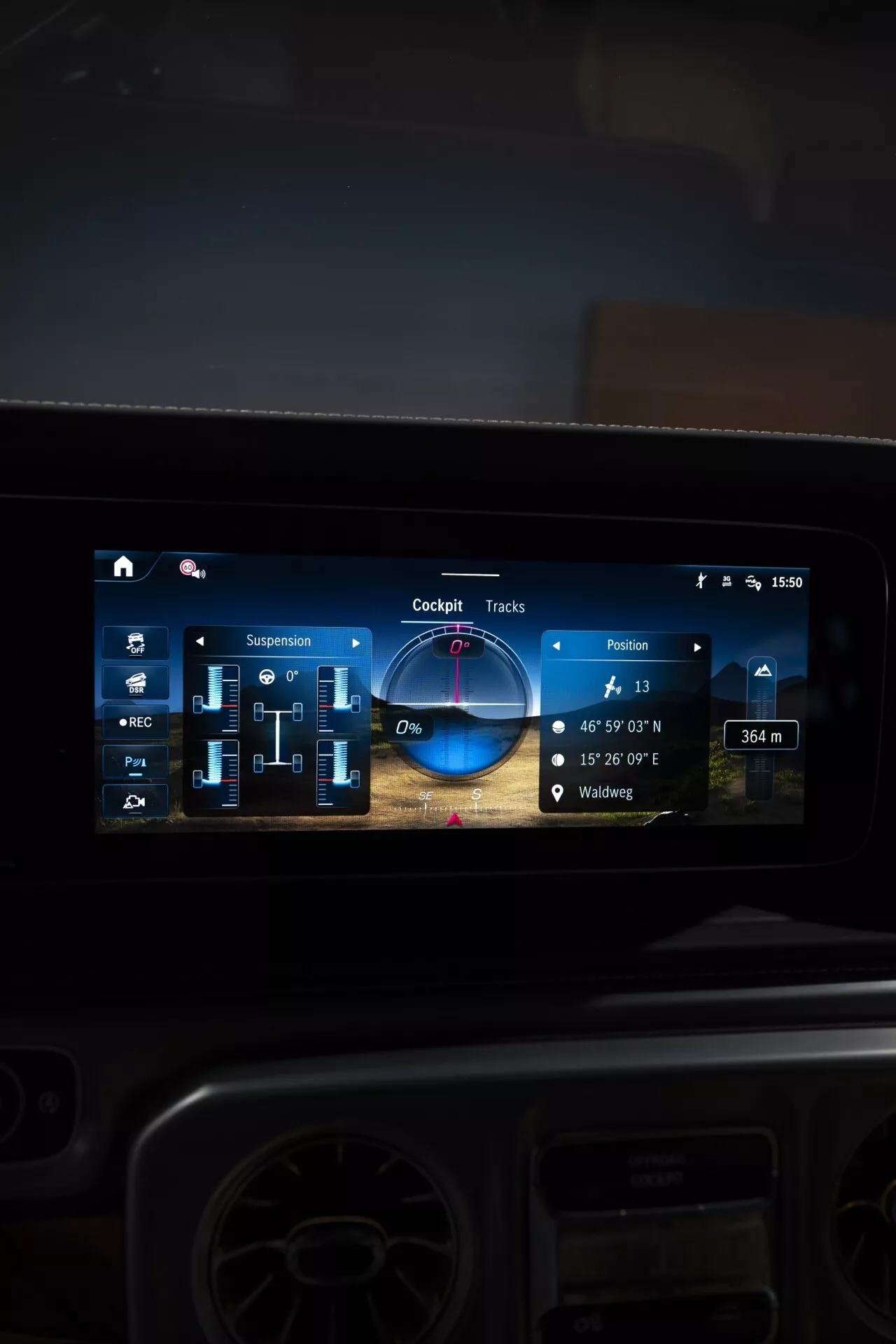 Zbliżenie na nowoczesny ekran dotykowy systemu infotainment w Mercedes-Benz G-Klasse 2024, przedstawiający nawigację, ustawienia zawieszenia, trasy terenowe i sterowanie multimediami, odzwierciedlające zaawansowane technologie samochodowe i luksusowy design wnętrza.