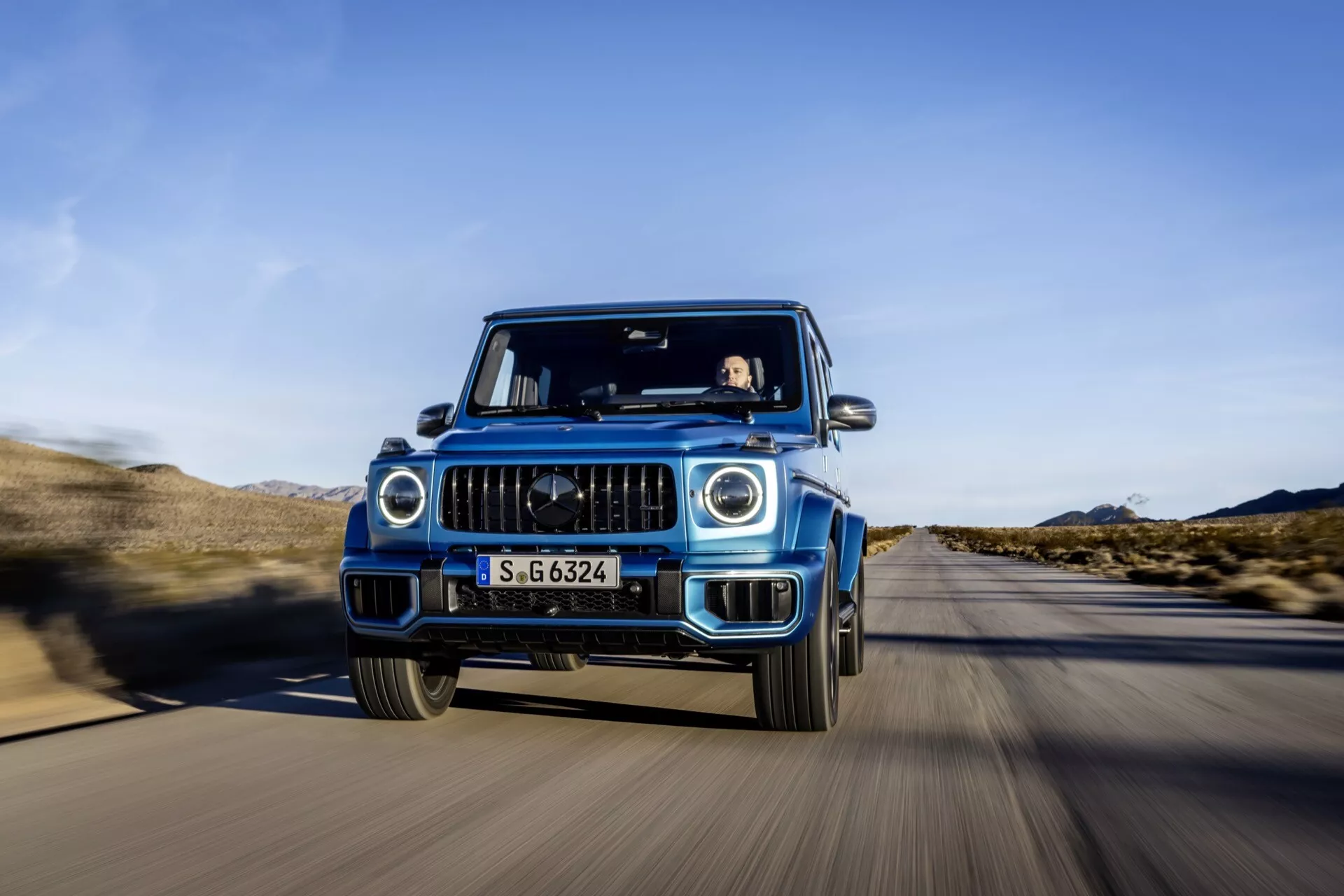 Luksusowy SUV Mercedes-Benz Klasy G 2024 w ruchu, uchwycony z przedniej perspektywy z zastosowaniem techniki śledzenia pokazującej szczegółową atrapę, reflektory LED i funkcje ADAS, na pustynnej drodze pod jasnoniebieskim niebem