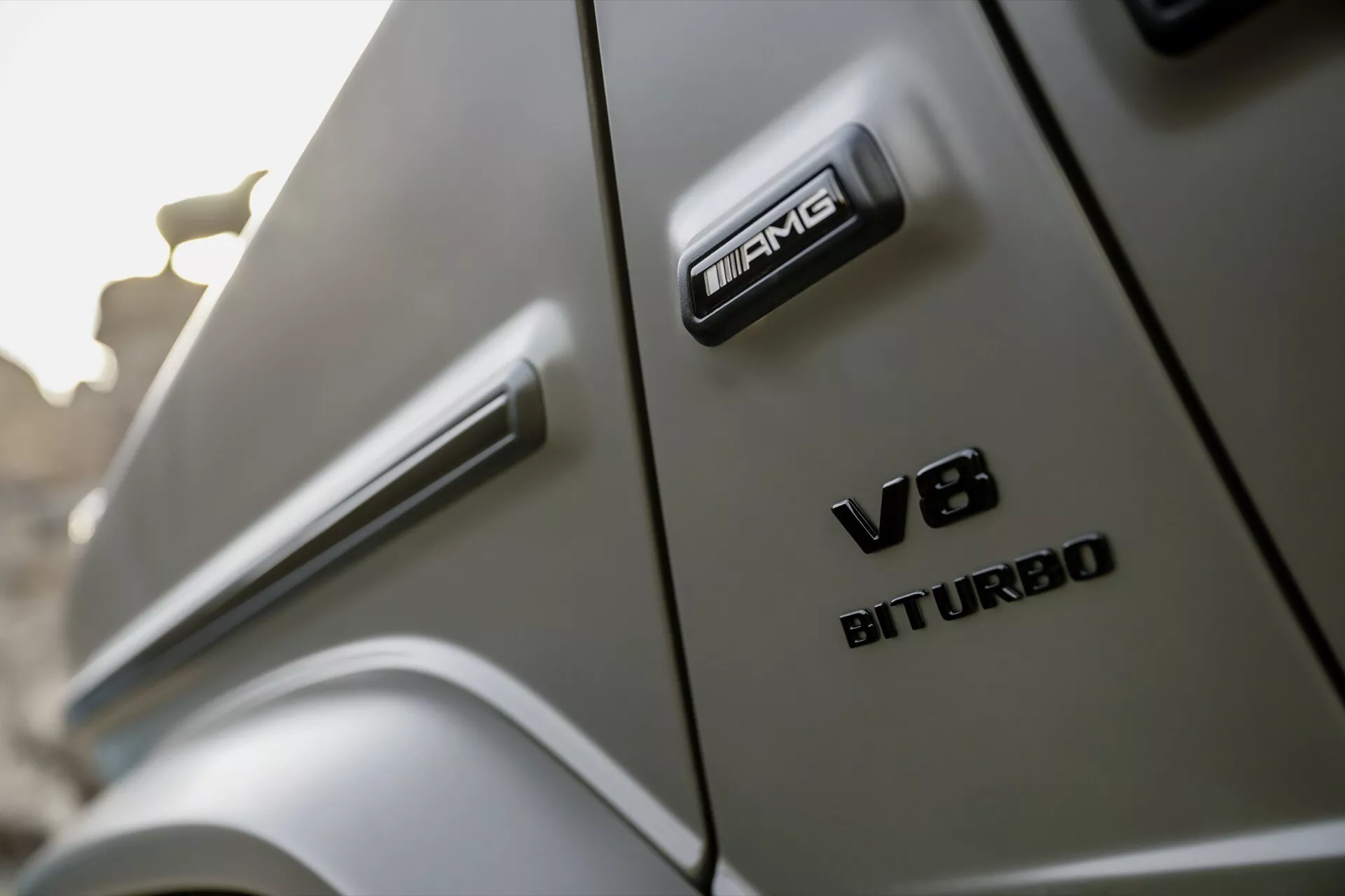Zbliżony widok pod kątem, prezentujący emblematy AMG i V8 BITURBO na nowym Mercedes-Benz G-Klasse 2024, podkreślający elementy designu wysokiej wydajności i luksusu pojazdu.
