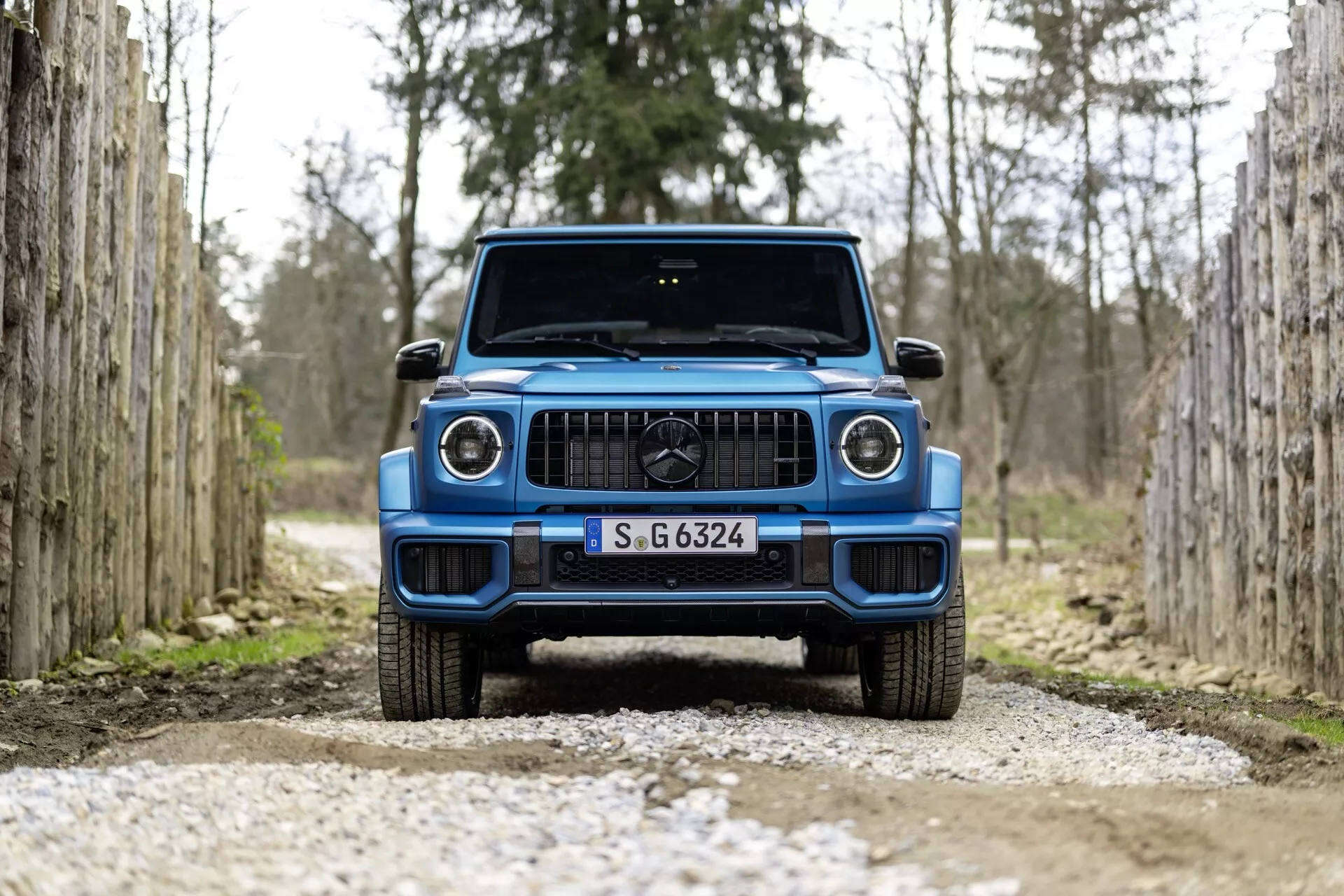 Frontalny widok niebieskiego SUV-a Mercedes-Benz G-Klasse 2024 na szutrowej ścieżce, pomiędzy rzędami drewnianych słupków, podkreślający nowoczesny design pojazdu i reflektory LED.