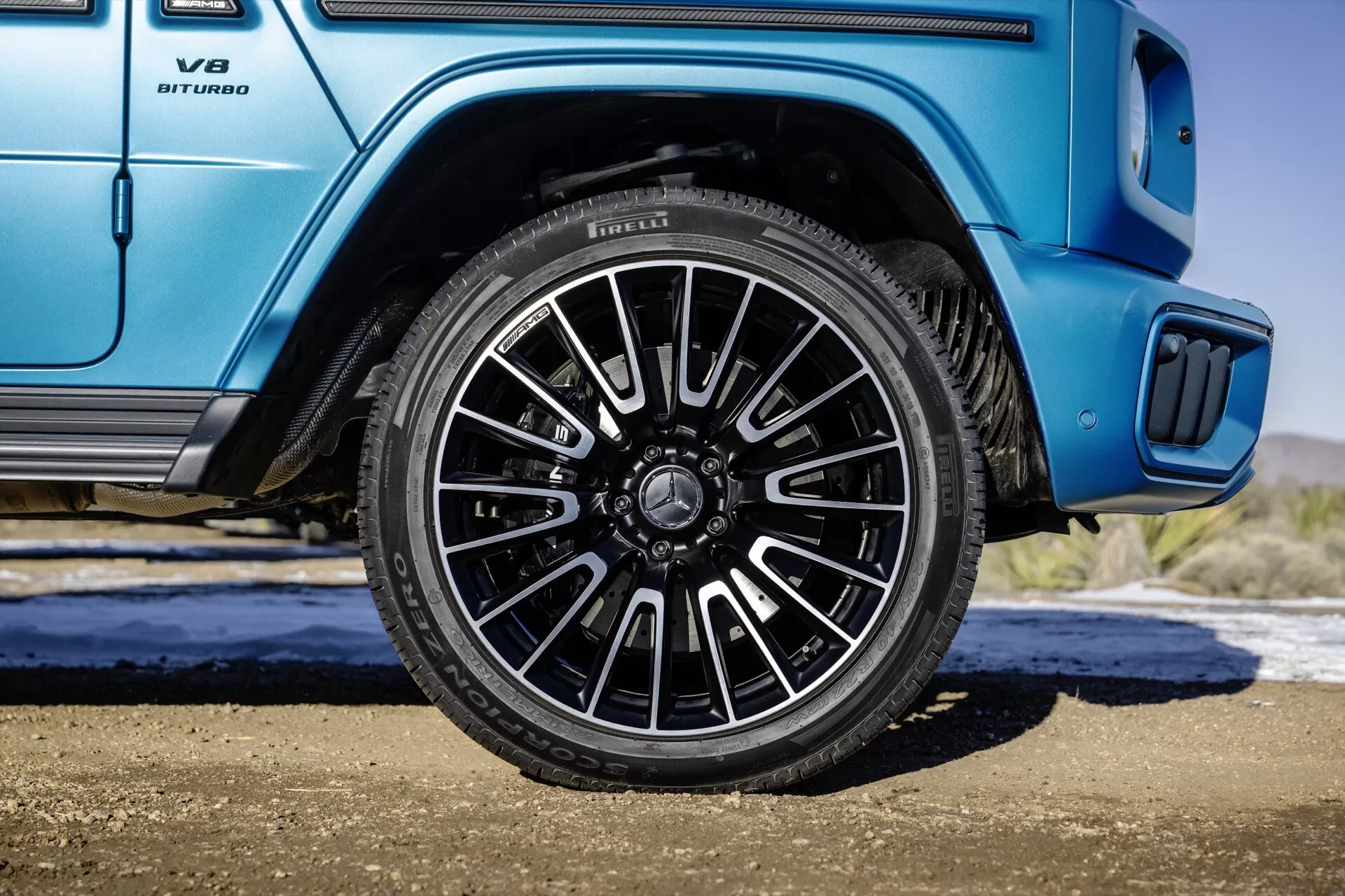 Mercedes-Benz Klasy G (G 465) na rok 2024 pokazuje koło od strony pasażera tyłu z wieloramieniowym wzorem i ciemnym wykończeniem na tle żywego niebieskiego nadwozia, w tym zbliżenie na emblemat V8 BITURBO i detal stopnia bocznego