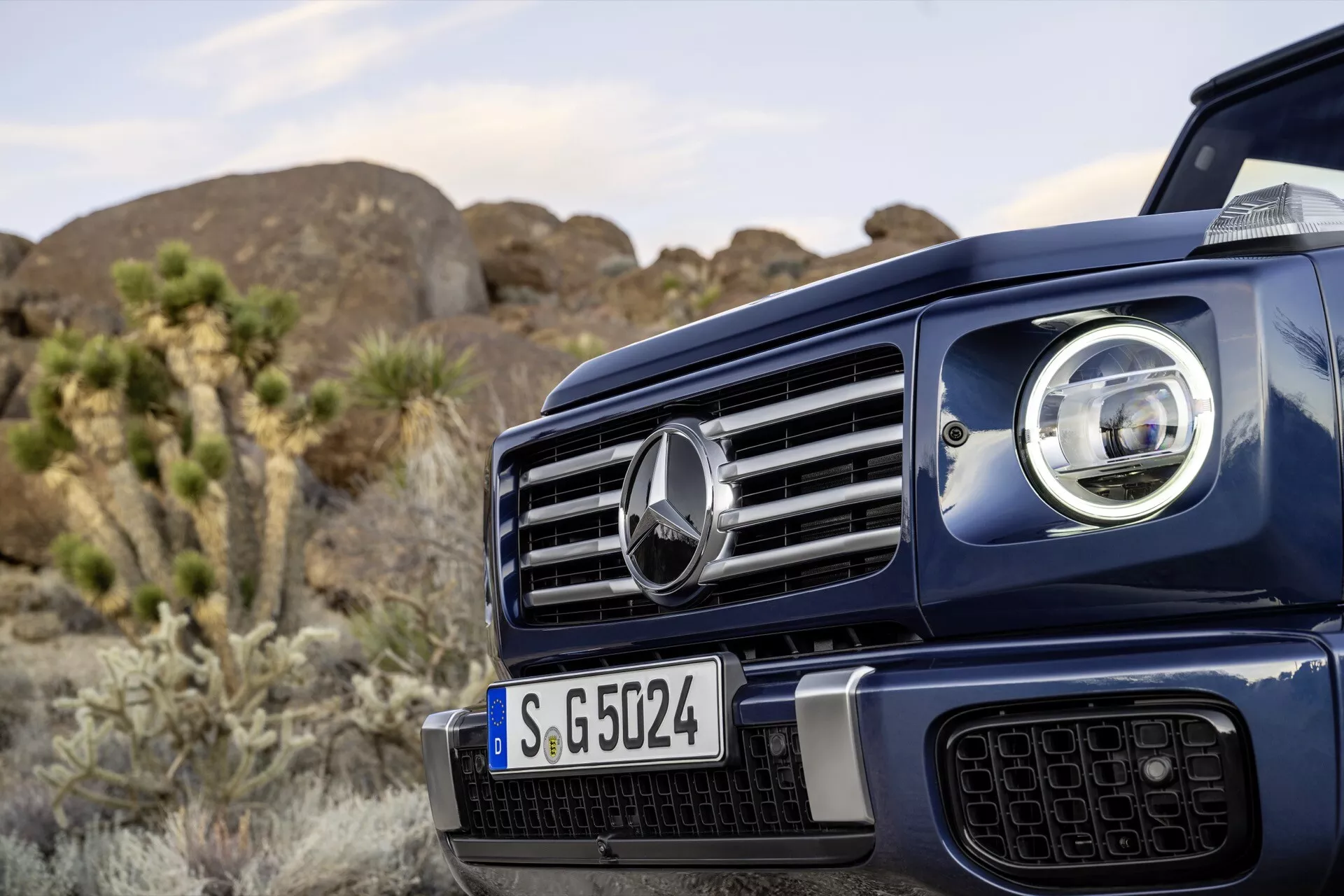 Zbliżony widok na przednią lewą stronę Mercedes-Benz G-Klasse 2024 z wyróżniającą się kratką z centralnym emblematem, częścią zaawansowanego reflektora LED, na tle naturalnego krajobrazu na zewnątrz sugerującym solidne zdolności terenowe