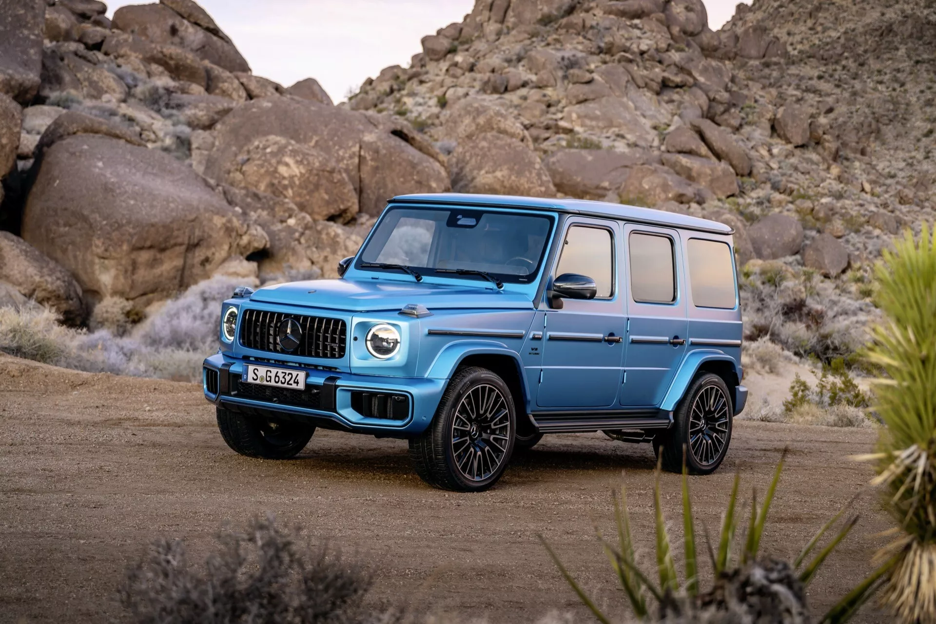 Luksusowy SUV Mercedes-Benz G-Klasse 2024 w kolorze jasnoniebieskim, prezentuje charakterystyczną atrapę z pionowymi listwami, duże wloty powietrza i reflektory LED z perspektywy trzy czwarte z przodu na nieutwardzonej nawierzchni z kamienną pustynią w tle, podkreślając jego zdolności terenowe i nowoczesne cechy stylistyczne.