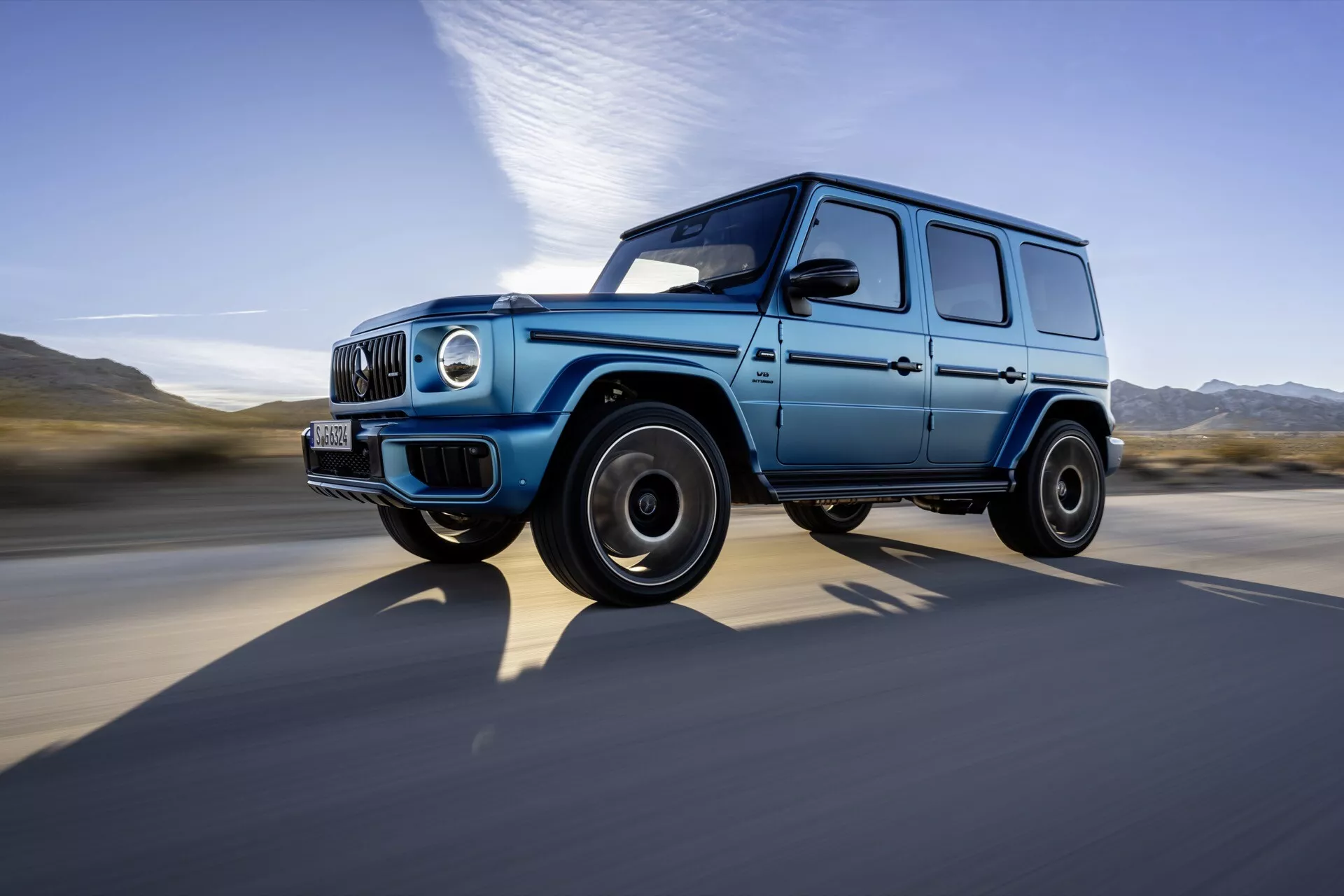 Luksusowy SUV Mercedes-Benz Klasy G z 2024 roku uchwycony w ruchu z dynamicznej perspektywy dolnej, pokazujący jego kanciaste, wytrzymałe wzornictwo, niebieski lakier z czarnymi elementami, okrągłe reflektory, diodowe światła dzienne i czarne koła ze stopów lekkich na tle rozmytym przez wysoką prędkość.