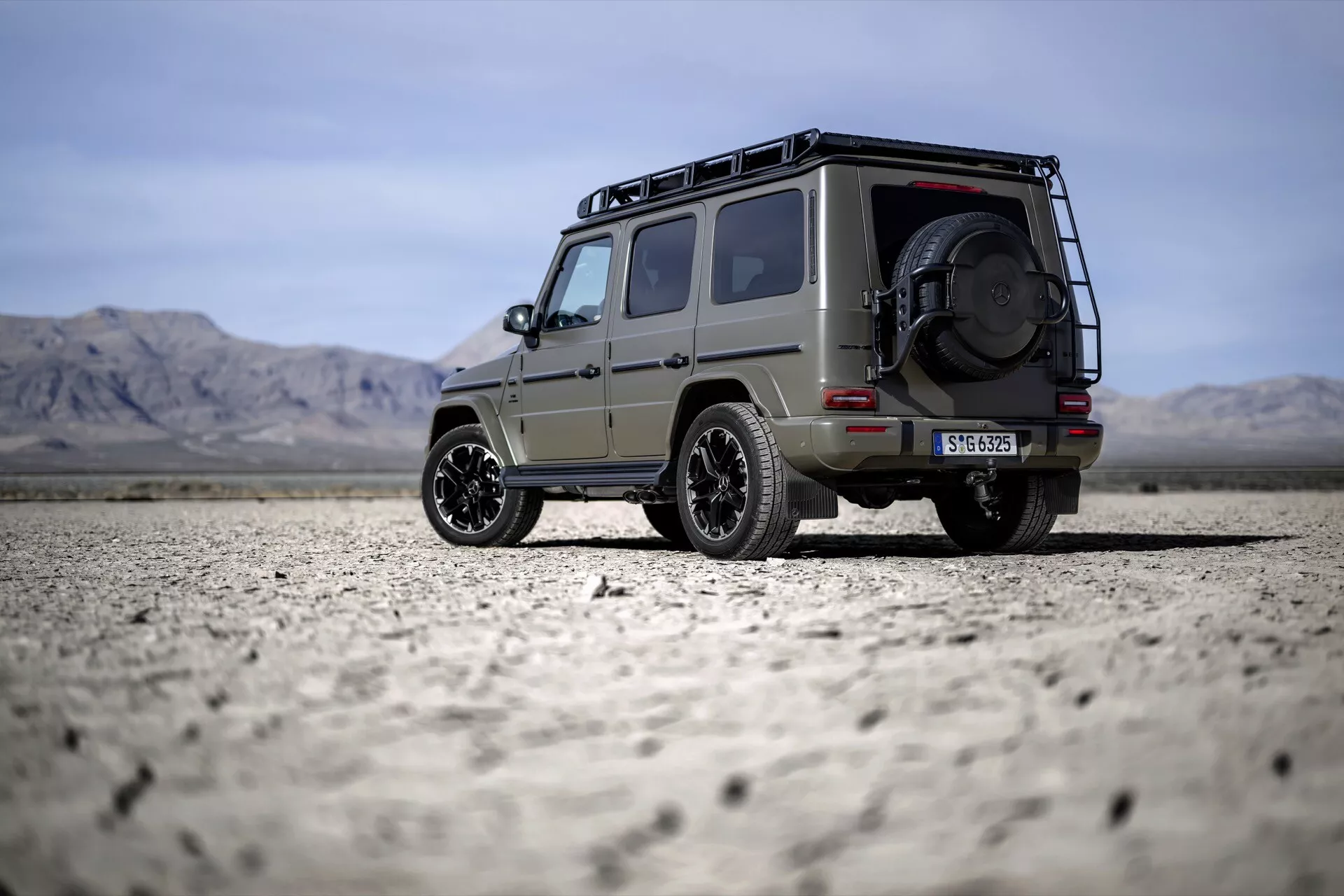 SUV Mercedes-Benz Klasy G 2024 zaparkowany na suchym dnie jeziora, pokazujący jego solidną konstrukcję z bagażnikiem dachowym, zapasowym kołem zamontowanym z tyłu i stylowymi felgami aluminiowymi pod czystym niebem z dalekimi górami.