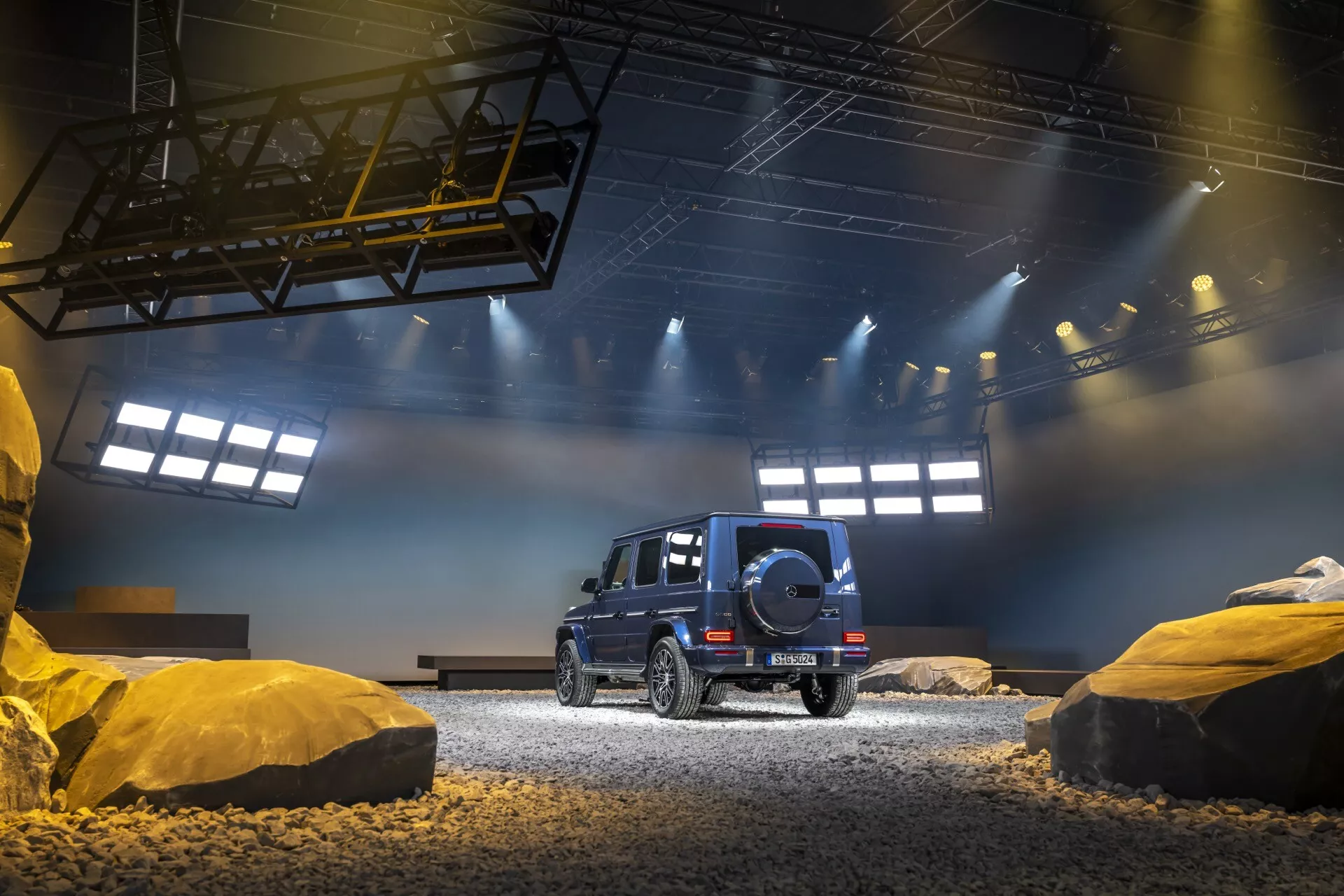 Czarne SUV Mercedes-Benz Klasy G (G-Klasse 465) rocznik 2024, widok z tyłu i boku, demonstrujące zdolności terenowe z zapasowym kołem, tylnymi światłami LED i luksusowym wzornictwem w inscenizowanym wnętrzu imitującym teren zwirowy z konstrukcjami podobnymi do skał pod dramatycznym sztucznym oświetleniem