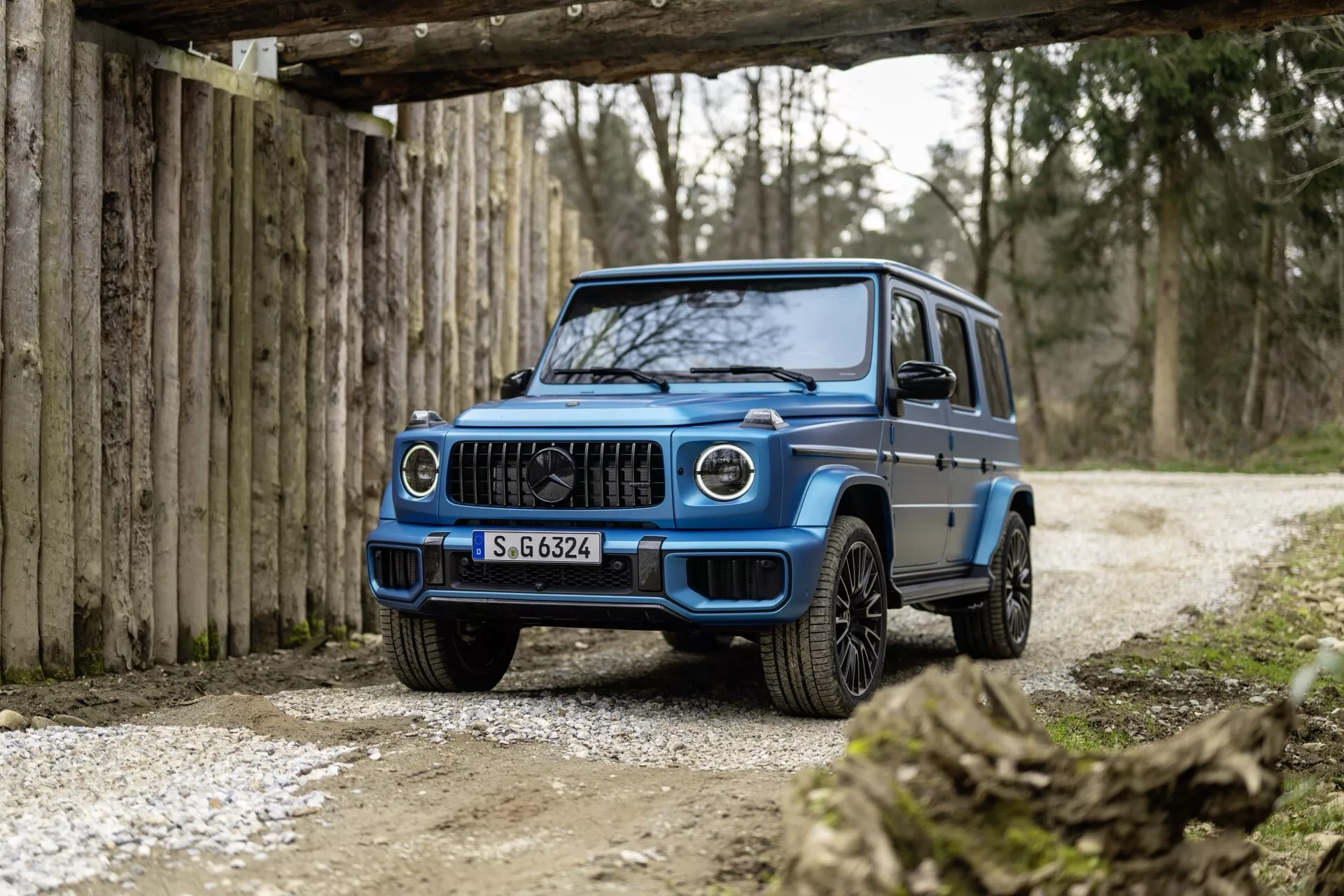 Trzy czwarte widoku z przodu na SUV Mercedes-Benz Klasy G 2024 w kolorze niebieskim, podkreślający grill przedni, reflektory LED, przedni zderzak i felgi aluminiowe, na tle rustykalnej drogi terenowej z łagodnym oświetleniem.
