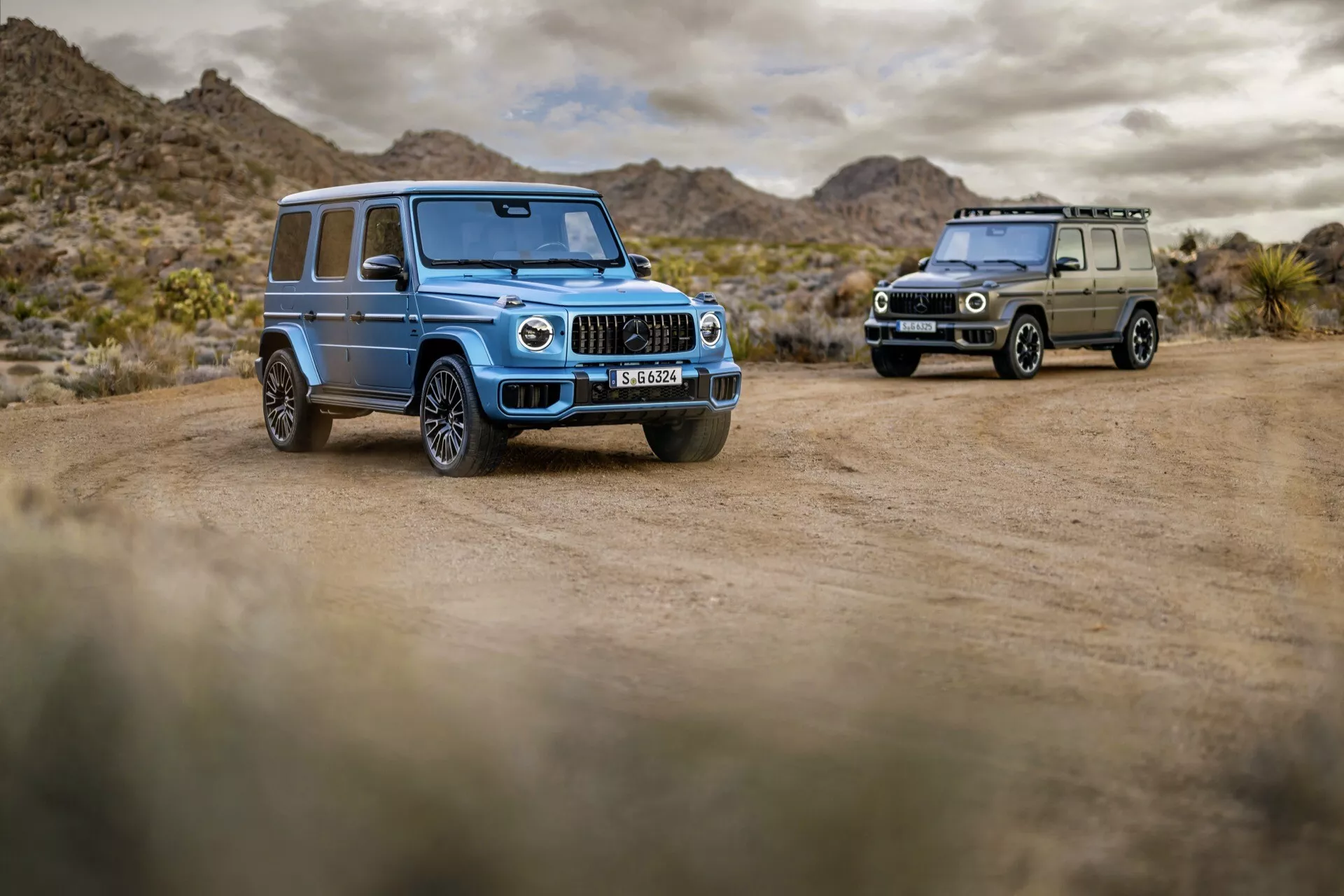 Mercedes-Benz Klasy G 2024 w jasnoniebieskim kolorze, wyeksponowany na pustynnym krajobrazie na drodze gruntowej, prezentujący reflektory LED, chromowaną grillę z pionowymi listwami, duże koła ze stopu z wieloma szprychami i opony terenowe, oraz zaawansowane funkcje technologiczne, z drugim samochodem Klasy G w kolorze oliwkowej zieleni widocznym w tle.