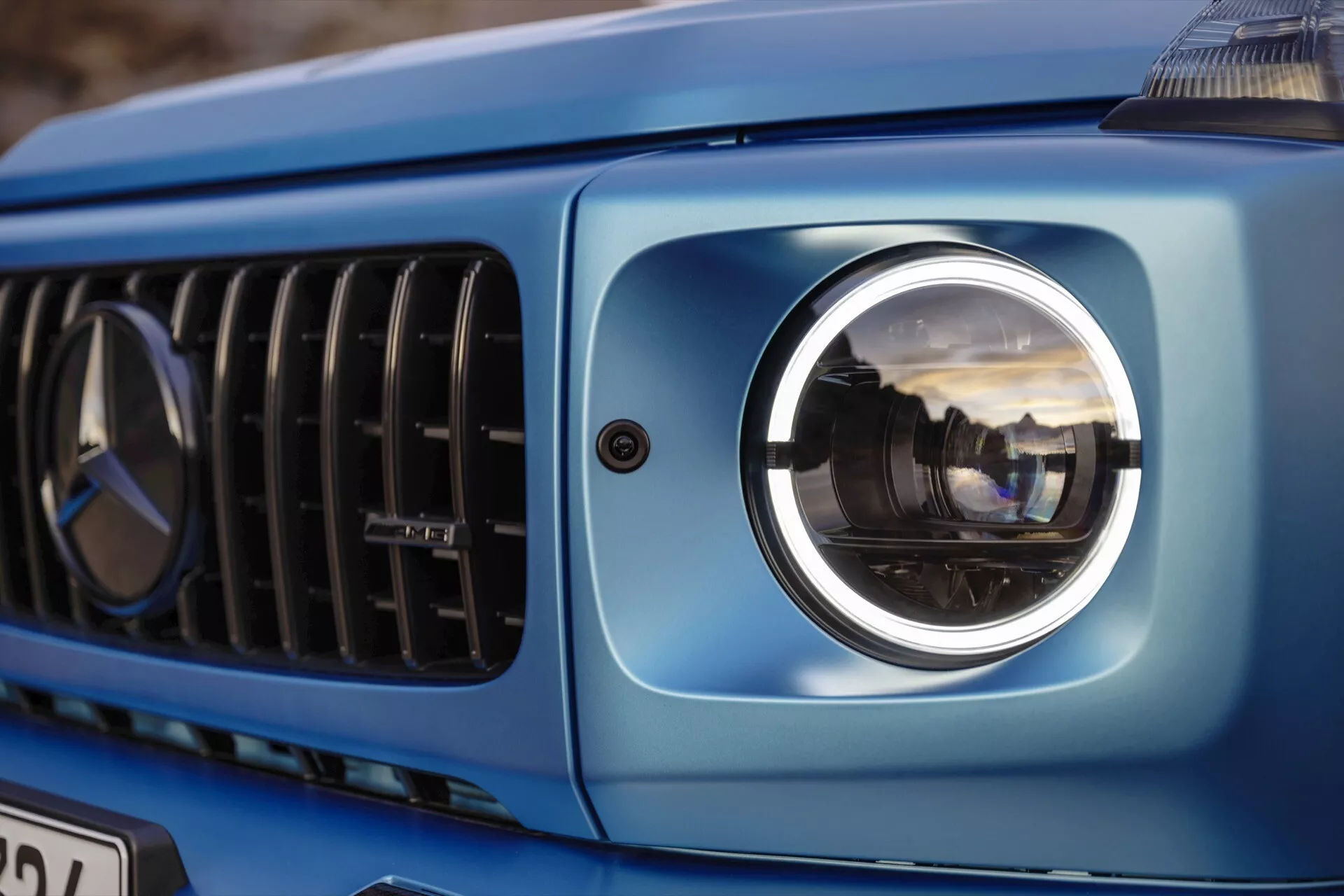 Zbliżenie na lewą przednią część Mercedes-Benz Klasy G 2024 ukazujące nowoczesny reflektor projektorowy, część grilla i metalicznie niebieską karoserię