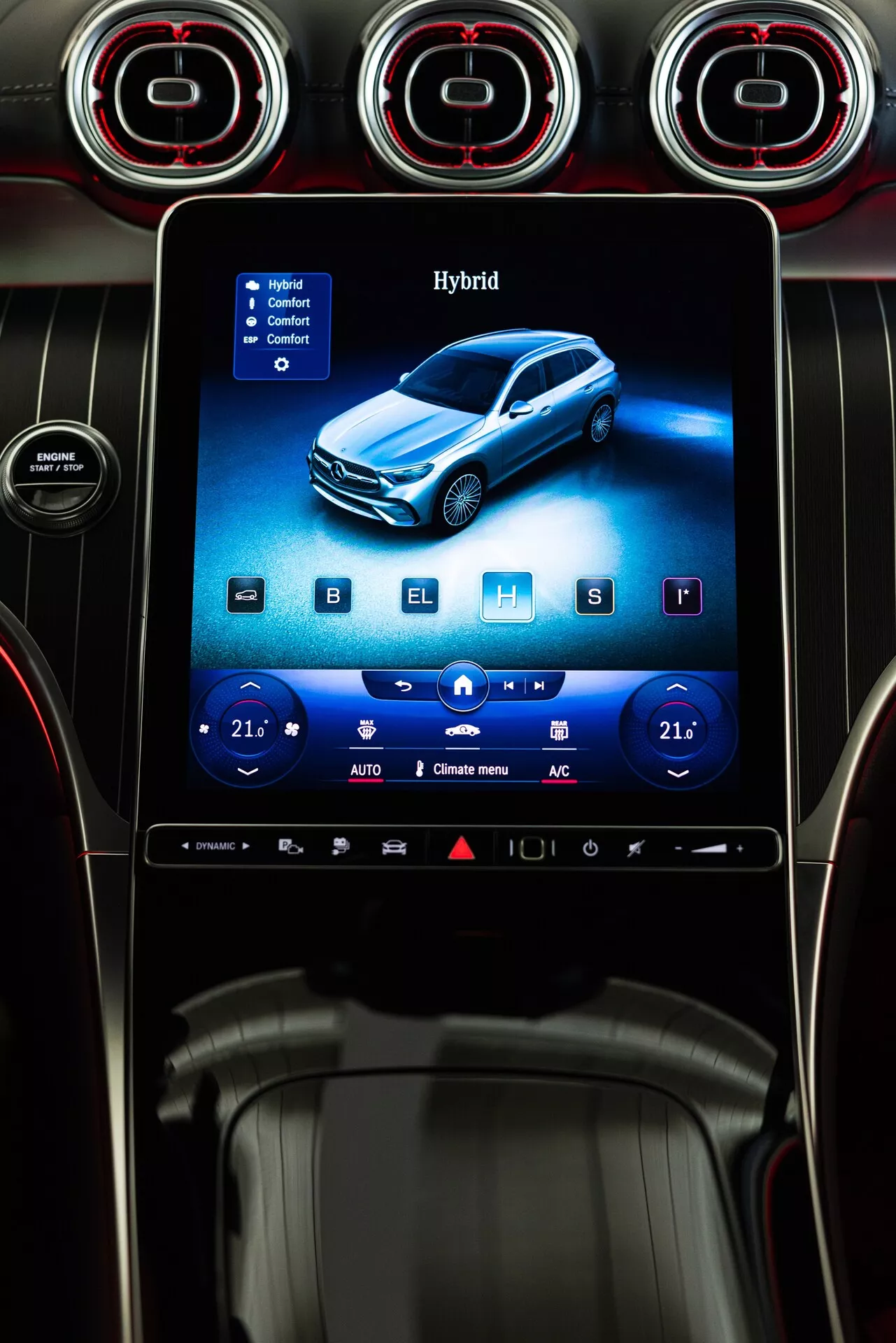 Zbliżenie na konsolę centralną Mercedes GLC 254 z 2022 roku, z pionowym systemem infotainment z ekranem dotykowym, otoczonym trzema okrągłymi wentylatorami z czerwonymi akcentami. Na ekranie widnieje niebieski pojazd hybrydowy, z cyfrowymi kontrolami klimatu ustawionymi na 21 stopni Celsjusza po obu stronach, sugerując podwójną kontrolę klimatu. Widoczne są ikony dla podgrzewania, chłodzenia siedzeń i opcji odmrażania, wraz z dolnym paskiem szybkiego dostępu. Konsola zaprojektowana jest z akcentami ze szczotkowanego metalu, błyszczącymi czarnymi powierzchniami i minimalistycznymi przyciskami, podkreślając nowoczesne i luksusowe wnętrze.