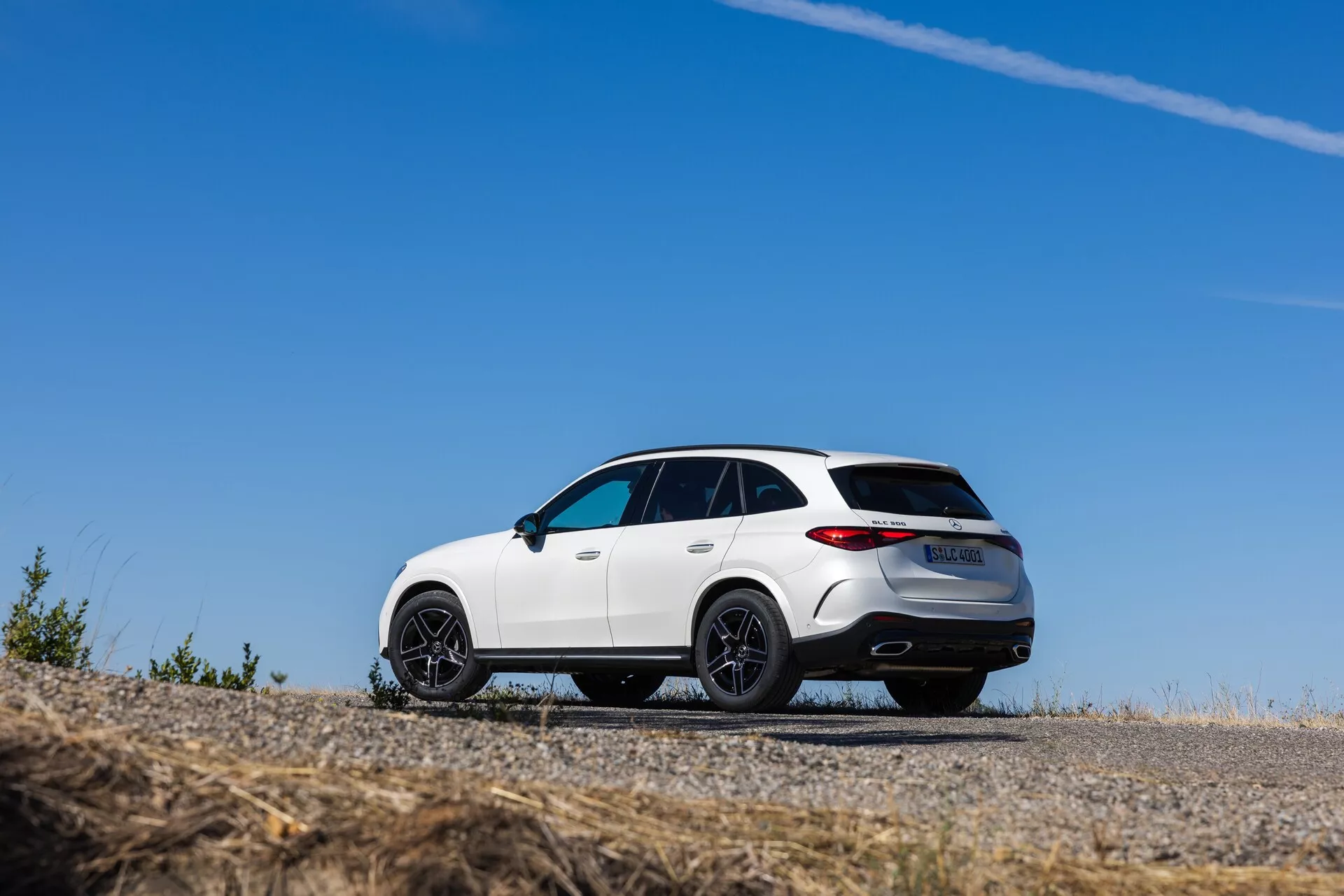 Niska perspektywa, trzech czwartych widoku tylu białego SUV-a Mercedes GLC 2022, zaparkowanego na asfaltowej powierzchni, prezentującego sportowy design z pochylonym dachem, dużymi czarnymi obręczami z wieloma szprychami i tylnymi światłami LED. Pojazd ma chromowane akcenty, podwójne końcówki wydechu, tylny spojler, panoramiczny dach i tylny dyfuzor, podkreślającą jego premium budowę i przywiązanie do osiągów.
