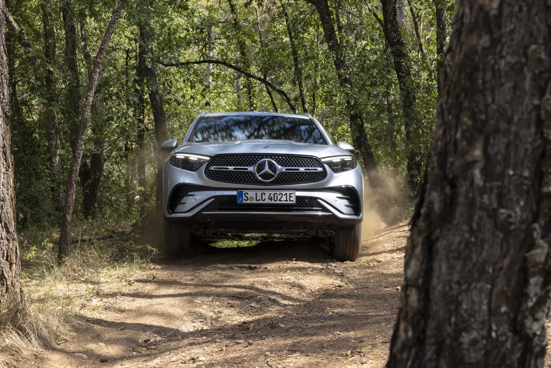 Widok z przodu na Mercedes GLC 2022 (254) na wąskiej, szutrowej drodze, otoczony drzewami, z wyłączonymi światłami, kurz za nim, słoneczny dzień z naturalnym światłem.