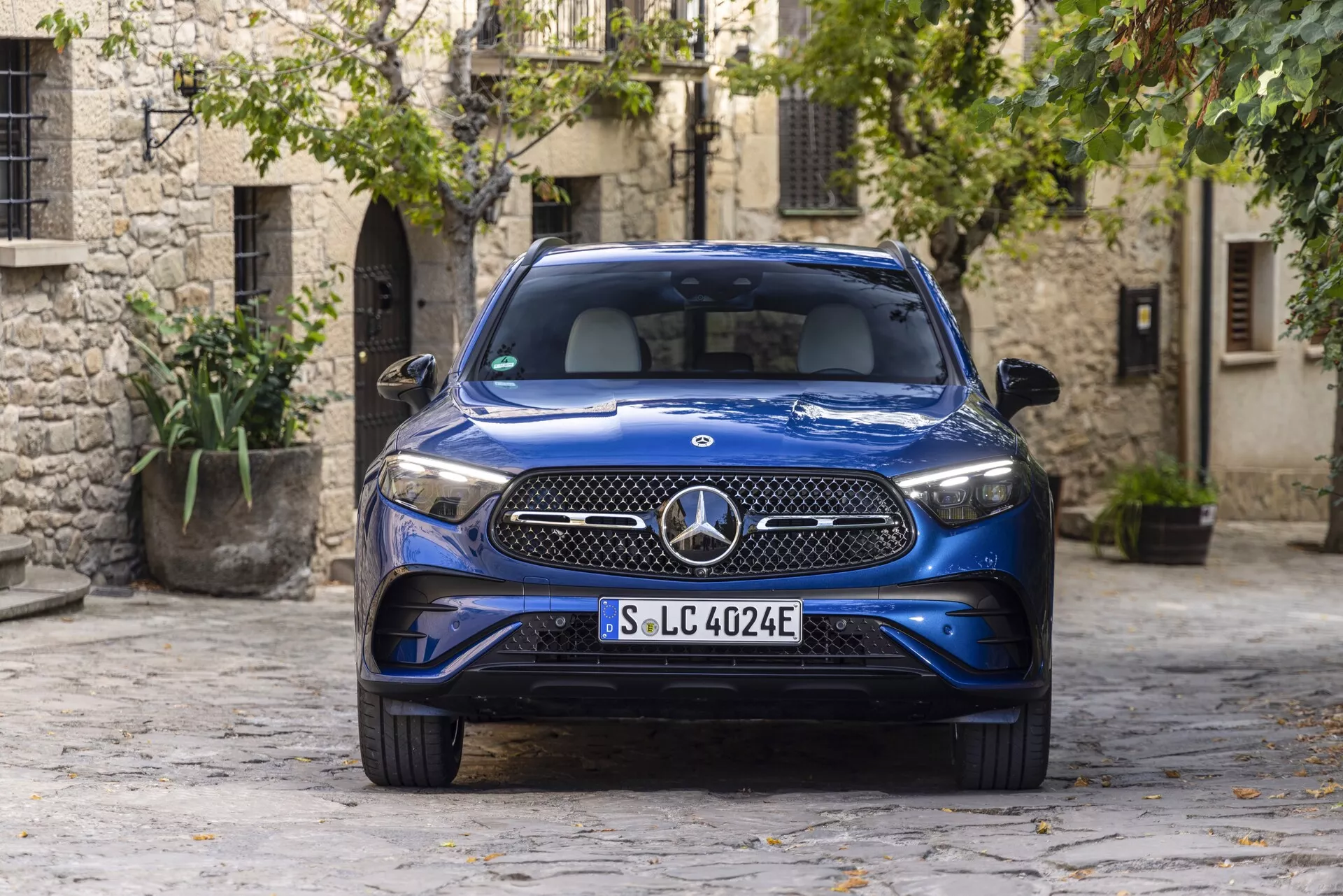 2022 Niebieski luksusowy SUV Mercedes GLC 254 zaparkowany w malowniczym brukowanym dziedzińcu starówki, prezentujący reflektory LED, charakterystyczny grille z dużym emblematem Mercedes-Benz, oznaczenie EQ wskazujące na elektryczną lub hybrydową przyrodę typu plug-in, elegancki metaliczny wygląd i zintegrowane wloty powietrza w przednim zderzaku. Ujęcie z kąta przedniego, podkreślając nowoczesny design motoryzacyjny na tle tradycyjnych kamiennych budynków i zieleni.