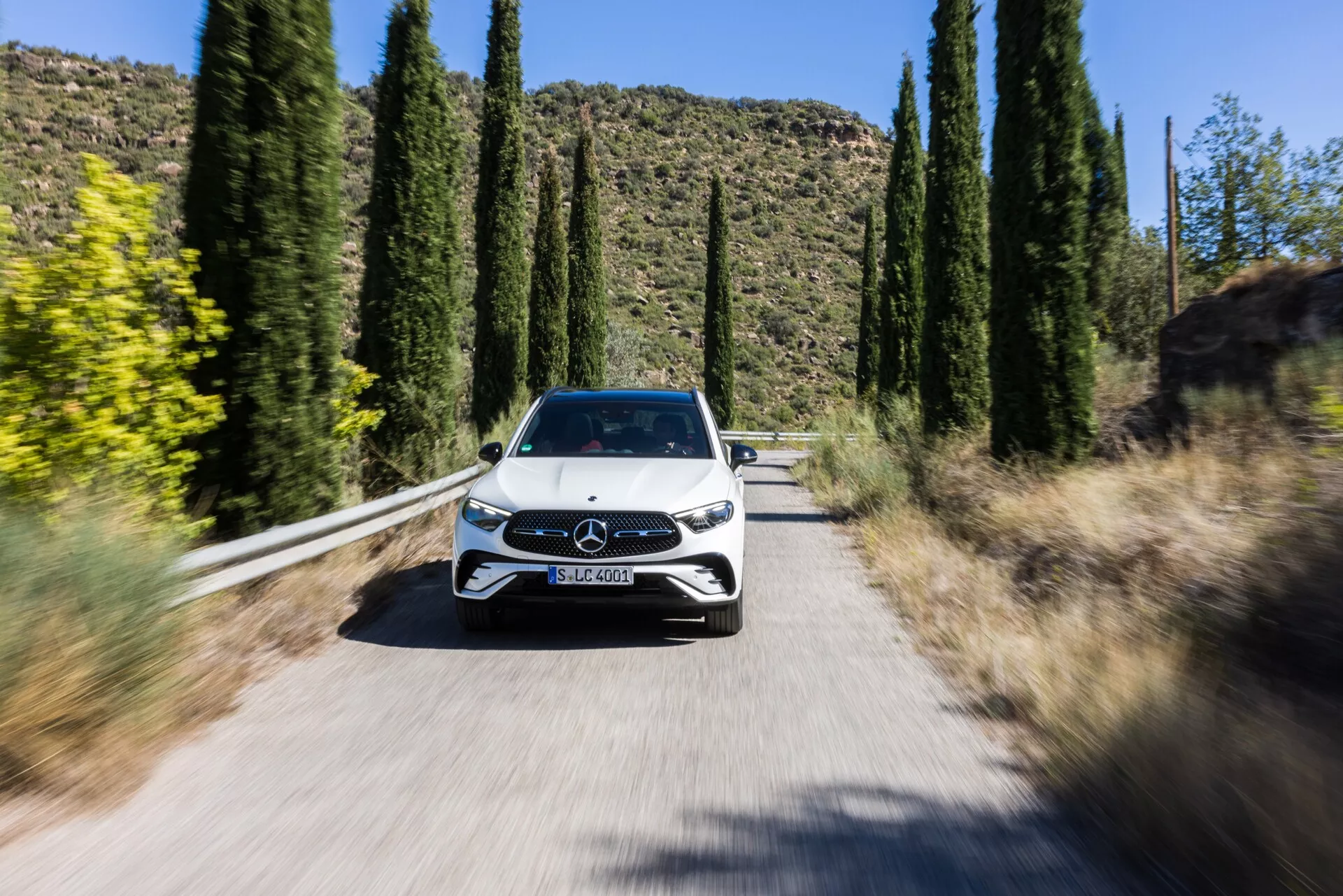 Dynamiczny obraz samochodu z perspektywy frontowej, Mercedes GLC 2022 (254) jadącego w kierunku kamery na wąskiej, nasłonecznionej drodze otoczonej wysokimi drzewami, ukazujący pełny przód z grillem z emblematem Mercedes-Benz, eleganckimi reflektorami LED, maską, przednią szybą i częścią dachu. Lekki rozmaz kół i rozmyte tło utworzone techniką rozmycia ruchu podkreślają ruch samochodu oraz zaawansowane funkcje, takie jak możliwa technologia reflektorów LED, zaktualizowany design grilla z aktywnymi elementami aerodynamicznymi lub czujniki do systemów wspomagania kierowcy, a także prawdopodobnie czujniki parkowania lub kamery.