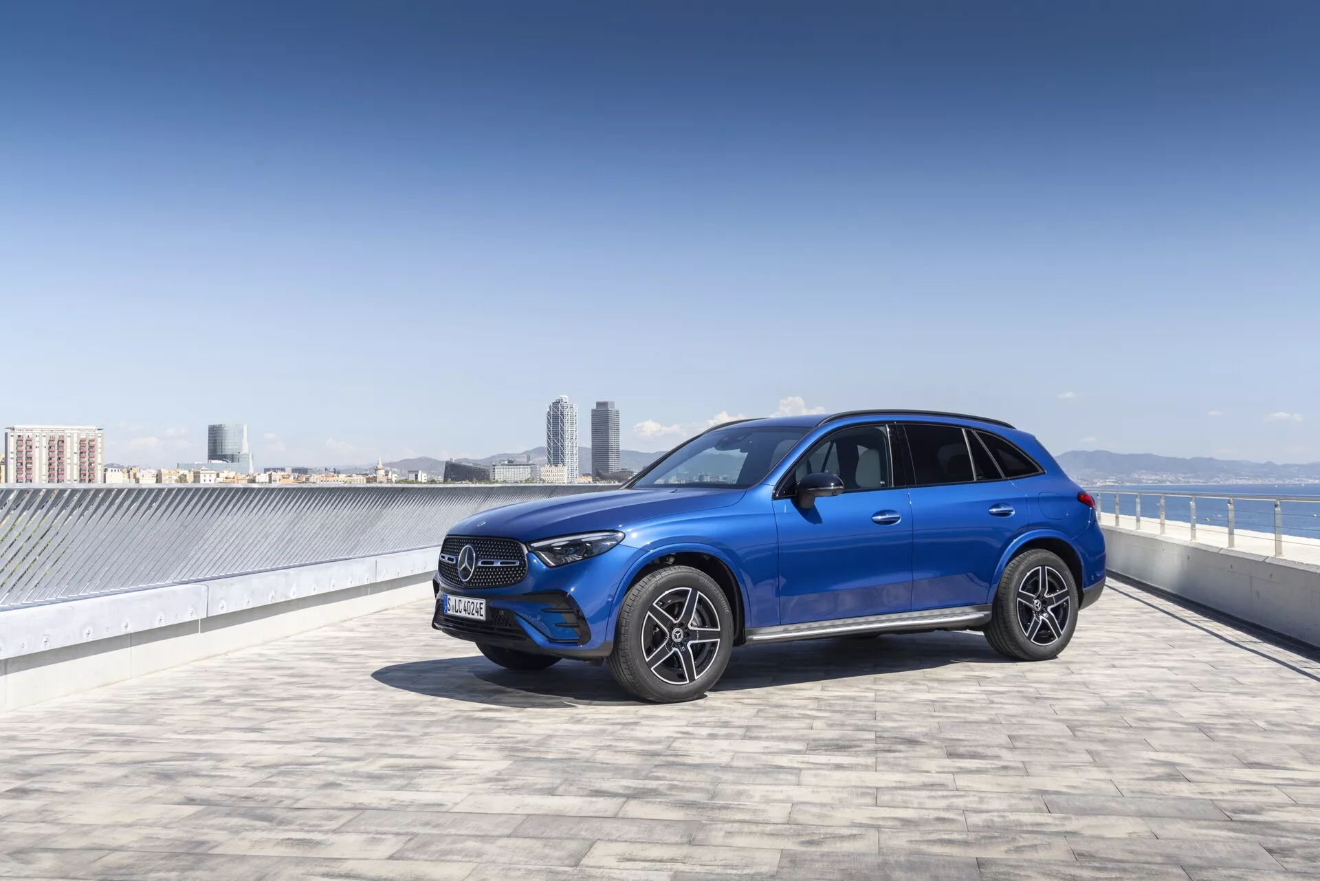Jasnoniebieski SUV Mercedes GLC 2022 na parkingu na dachu z czystym niebem i miejską panoramą w tle, uchwycony z przodu w trzy czwarte. Obraz podkreśla wyraźny grill przedni z dużym emblematem, kątowe reflektory LED, przedni zderzak z możliwymi czujnikami, wieloszprychowe felgi aluminiowe i lusterka boczne z zintegrowanymi kierunkowskazami. Smukłe linie i sportowa postawa samochodu są dobrze podkreślone, sugerując zaawansowaną technologię i miejską elegancję.