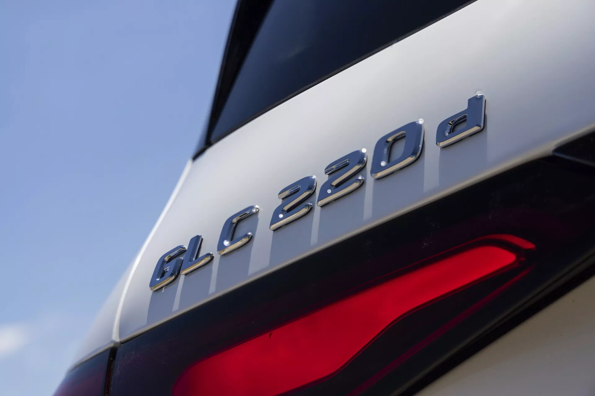 Zbliżenie tylnej części Mercedes-Benz GLC 220d 2022, z wyróżnionym oznaczeniem modelu tuż nad częściowo widocznym tylnym światłem. Obraz skupia się na oznaczeniu 'GLC 220d', sugerując, że jest to wariant diesla. Zdjęcie z niskiego, lekko podnoszącego się kąta ukazuje eleganckie linie projektu, metaliczne wykończenie oznaczenia kontrastujące z błyszczącym lakierem oraz nowoczesne elementy LED tylnego światła, wskazujące na zaawansowaną technologię oświetleniową.