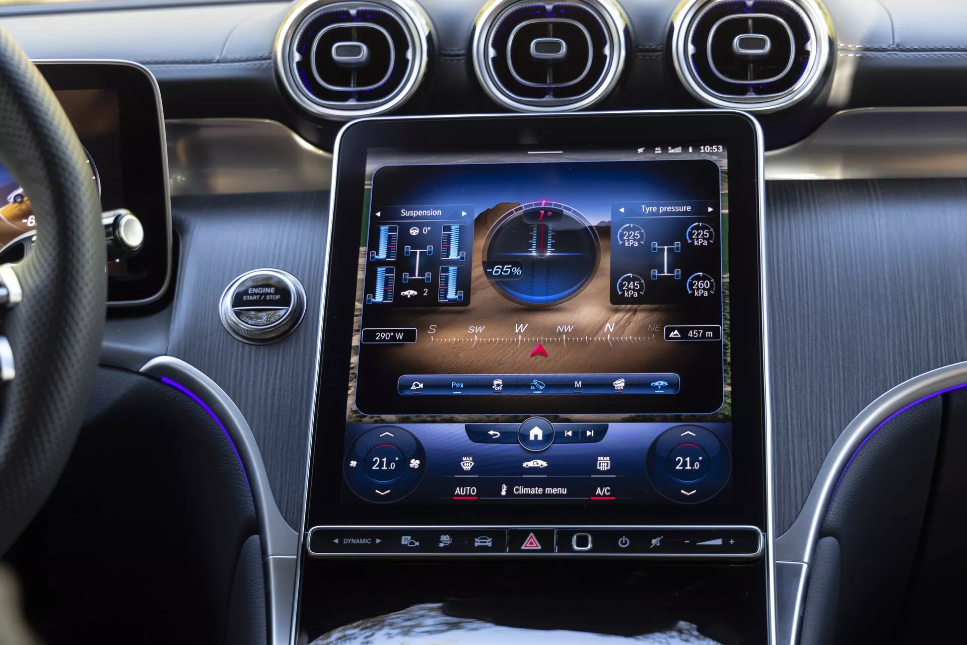 Widok wnętrza Mercedes GLC 2022 skupiający się na centralnym systemie infotainment i desce rozdzielczej, z dużym ekranem dotykowym o wysokiej rozdzielczości, w tym mapą nawigacji GPS, informacjami o ciśnieniu w oponach i wskaźnikiem ekologicznej jazdy. Poniżej ekranu znajdują się sterowniki klimatyzacji dwustrefowej, a powyżej trzy okrągłe wloty powietrza z metalicznym wykończeniem. Zdjęcie, wykonane z perspektywy pasażera, pokazuje również część kierownicy i sugeruje obecność wysokiej jakości materiałów we wnętrzu, takich jak szczotkowany metal i skóra.