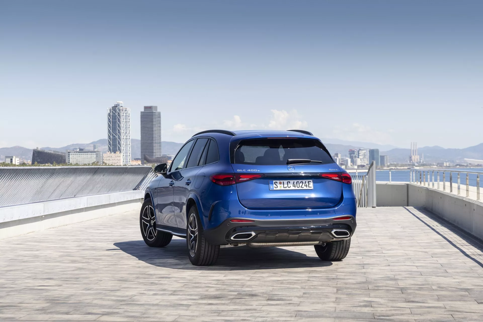 Zdjęcie Mercedesa GLC 2022 (254) z niskiej perspektywy, widok z tyłu z ukosa, ukazujące eleganckie i sportowe linie projektowe, tylne światła, aluminiowe felgi, metaliczny lakier i oznaczenia. Obraz ukazuje całą wysokość i szerokość pojazdu, podkreślając takie elementy jak tylna szyba, układ wydechowy, tablica rejestracyjna oraz potencjalnie emblematu wersji wyposażenia lub zespołu napędowego. Sceneria na zewnątrz, prawdopodobnie na dachu parkingu, z miejską linią horyzontu w tle, podkreśla nowoczesny i wyrafinowany wygląd pojazdu.