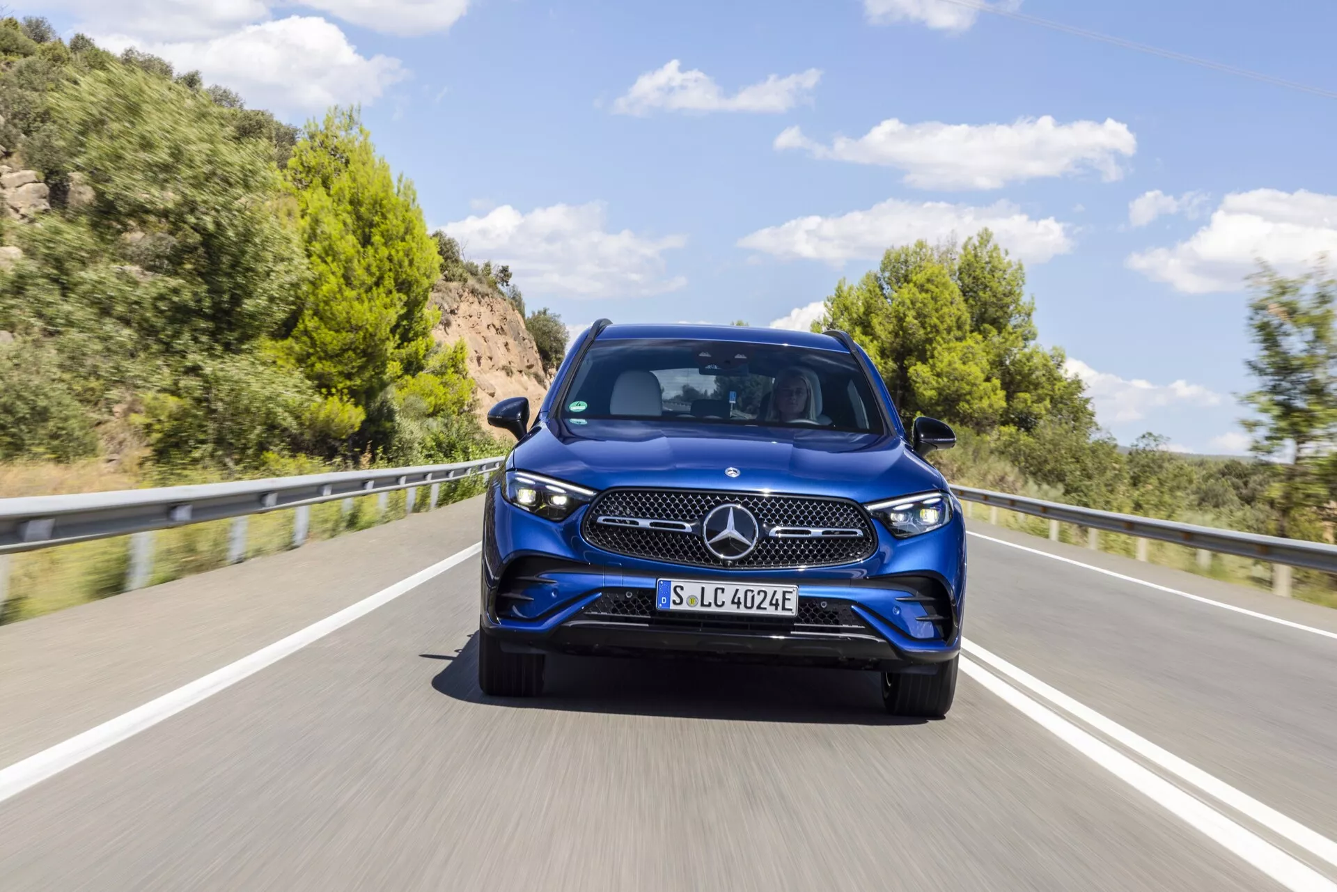 Obraz niebieskiego modelu Mercedes-Benz GLC 254 z 2022 roku uchwyconego pod kątem trzech czwartych przodu, podkreślający elegancki design, wyraźny trójramienny emblemat, szczegółowy wzór grilla, eleganckie reflektory wtapiające się w błotniki i wyraźne detale przedniego zderzaka, maski i drzwi. Samochód, sfotografowany w ruchu na malowniczej dwupasmowej drodze z zielonymi wzgórzami w tle, prawdopodobnie posiada technologię elektryczną lub hybrydową, co sugeruje oznaczenie 'EQ' na uchwycie tablicy rejestracyjnej. Obraz jest szczegółowy, z rozmyciem ruchu wskazującym ruch i pokazuje światła dzienne samochodu w ciągu dnia przy czystym niebie.