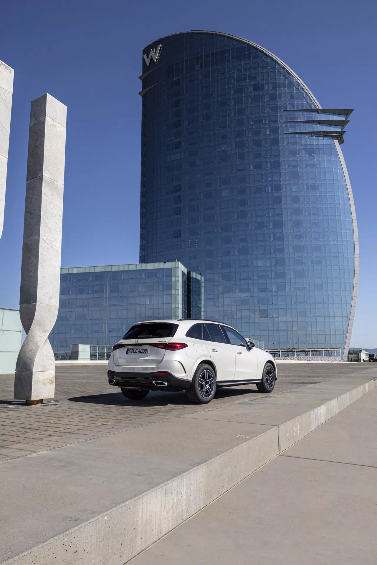 Mercedes GLC 2022 - Biały luksusowy SUV zaparkowany na placu; średnio-niski kąt uchwycenia profilu bocznego od strony pasażera i tyłu, pokazujący nowoczesne tylne światła, podwójny wydech, wieloszprychowe felgi aluminiowe, linię dachu typu coupé, przyciemniane szyby; w tle współczesny budynek hotelowy lub biurowy z odbijającą światło fasadą szklaną w jasnym świetle dziennym.