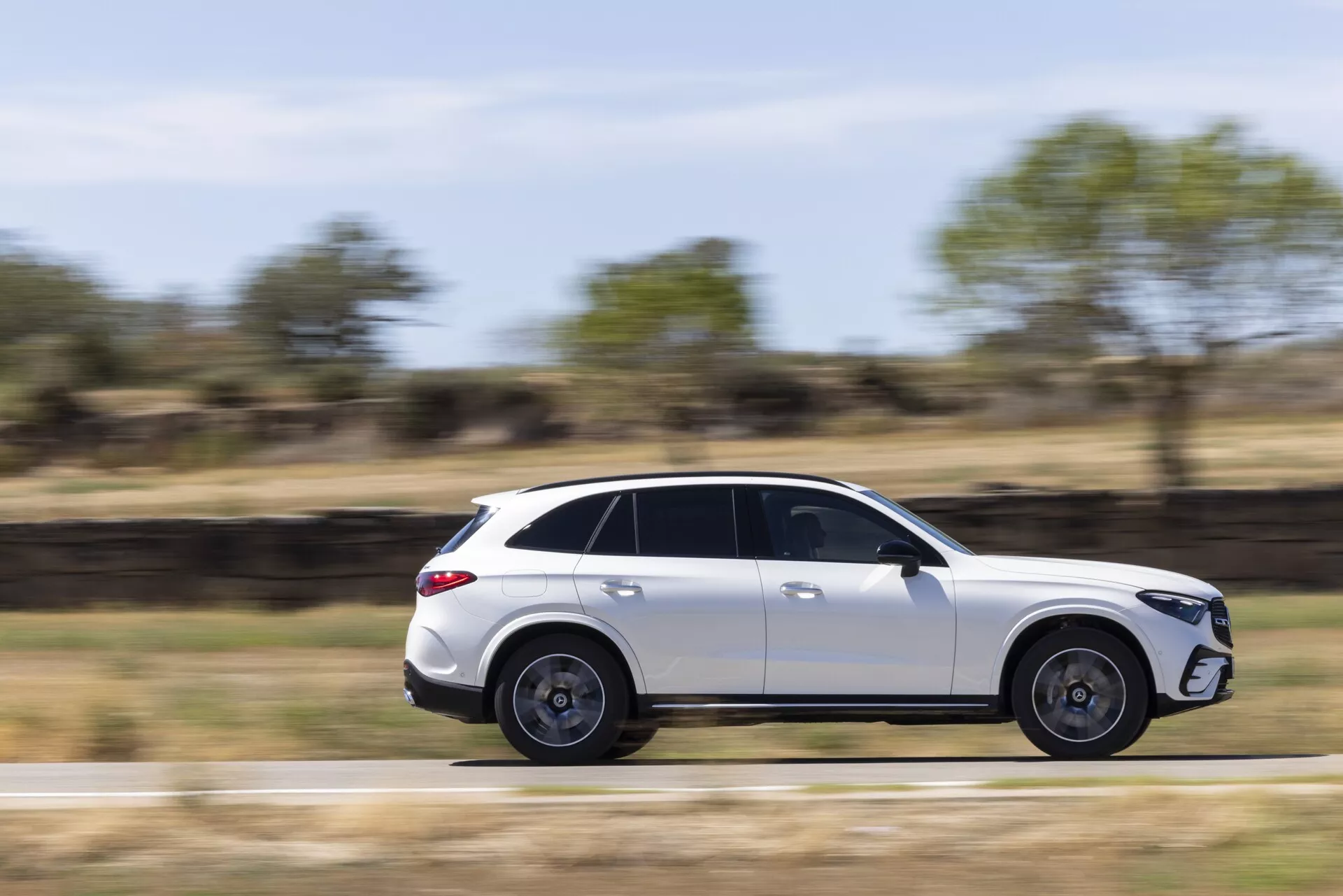 Obraz nowoczesnego, eleganckiego białego SUV-a Mercedes GLC 2022 w ruchu, widzianego z profilu bocznego. Pojazd został uchwycony przy użyciu techniki panoramowania, co utrzymuje go w centrum ostrości, jednocześnie rozmazując tło, aby pokazać ruch. Z przyciemnianymi szybami, dachem panoramicznym, pięcioramiennymi kołami aluminiowymi i aerodynamicznym designem, ten SUV jest ustawiony na tle wiejskim oświetlonym naturalnym światłem dziennym.
