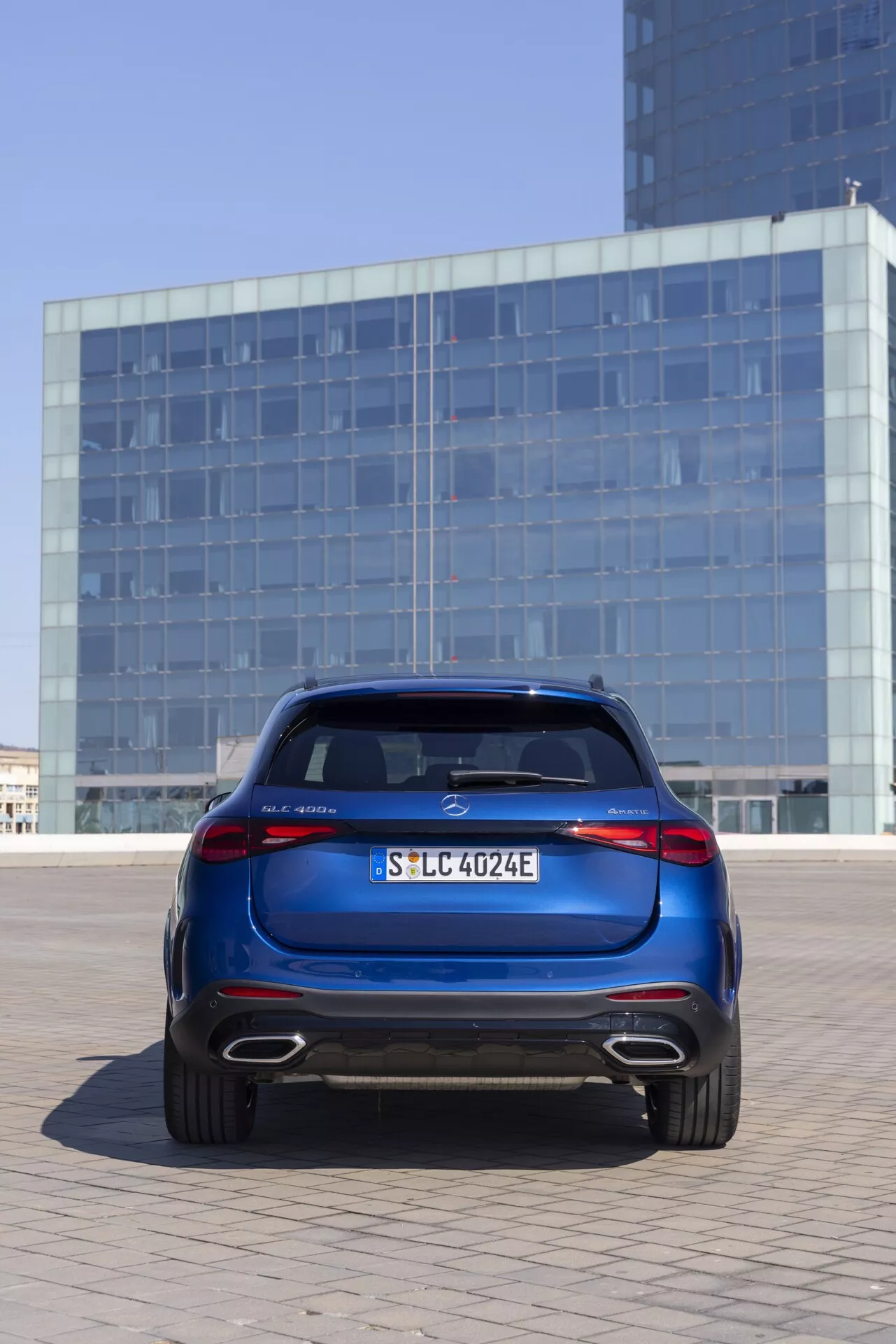 Widok z tyłu na Mercedes GLC 2022 (254) w głębokim metalicznym niebieskim kolorze, umieszczony na zewnątrz w miejskim otoczeniu z budynkiem o szklanej fasadzie w tle. Samochód jest umieszczony centralnie i uchwycony z nieco niskiego kąta, podkreślając pokrywę bagażnika, tylny zderzak, tylne światła LED, felgi aluminiowe i spojler dachowy. Cechy obejmują wyraźny centralny znaczek, tablicę rejestracyjną, oznaczenie modelu po lewej stronie, 'E' wskazujące na model elektryczny lub hybrydowy po prawej oraz tylny dyfuzor z wyjściami wydechowymi, sugerującymi model hybrydowy.