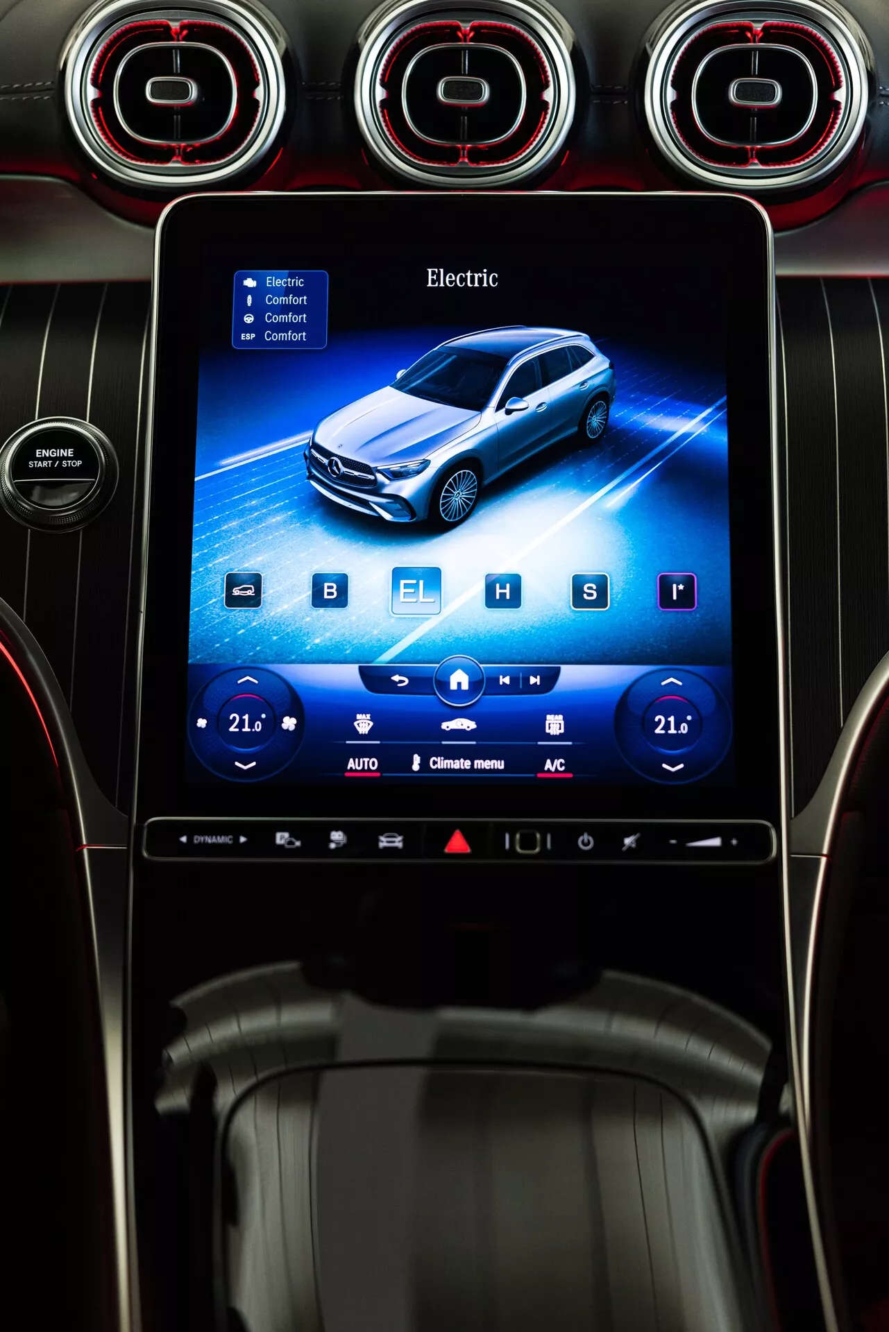 Widok wnętrza deski rozdzielczej samochodu z systemem infotainment Mercedes GLC 2022. Głównym punktem jest pionowo zorientowany ekran dotykowy wbudowany w deskę rozdzielczą. Żywy interfejs przedstawia graficzne zobrazowanie niebieskiego pojazdu elektrycznego w lekkiej 3/4 przedniej perspektywie. Na górze ekranu wyświetlane jest słowo 'Electric', wskazując na typ napędu pojazdu. Ekran zawiera również ustawienia klimatyzacji dwustrefowej z temperaturą ustawioną na '21' po obu stronach, wraz z przyciskami regulacji plus i minus oraz różnymi ikonami na dole. Materiały deski rozdzielczej otaczające ekran wydają się obejmować błyszczące i teksturowane tworzywa sztuczne oraz być może elementy skórzane. Powyżej ekranu znajdują się okrągłe otwory wentylacyjne z czerwonymi akcentami, dodając wnętrzu samochodu sportowy lub ekskluzywny wygląd.
