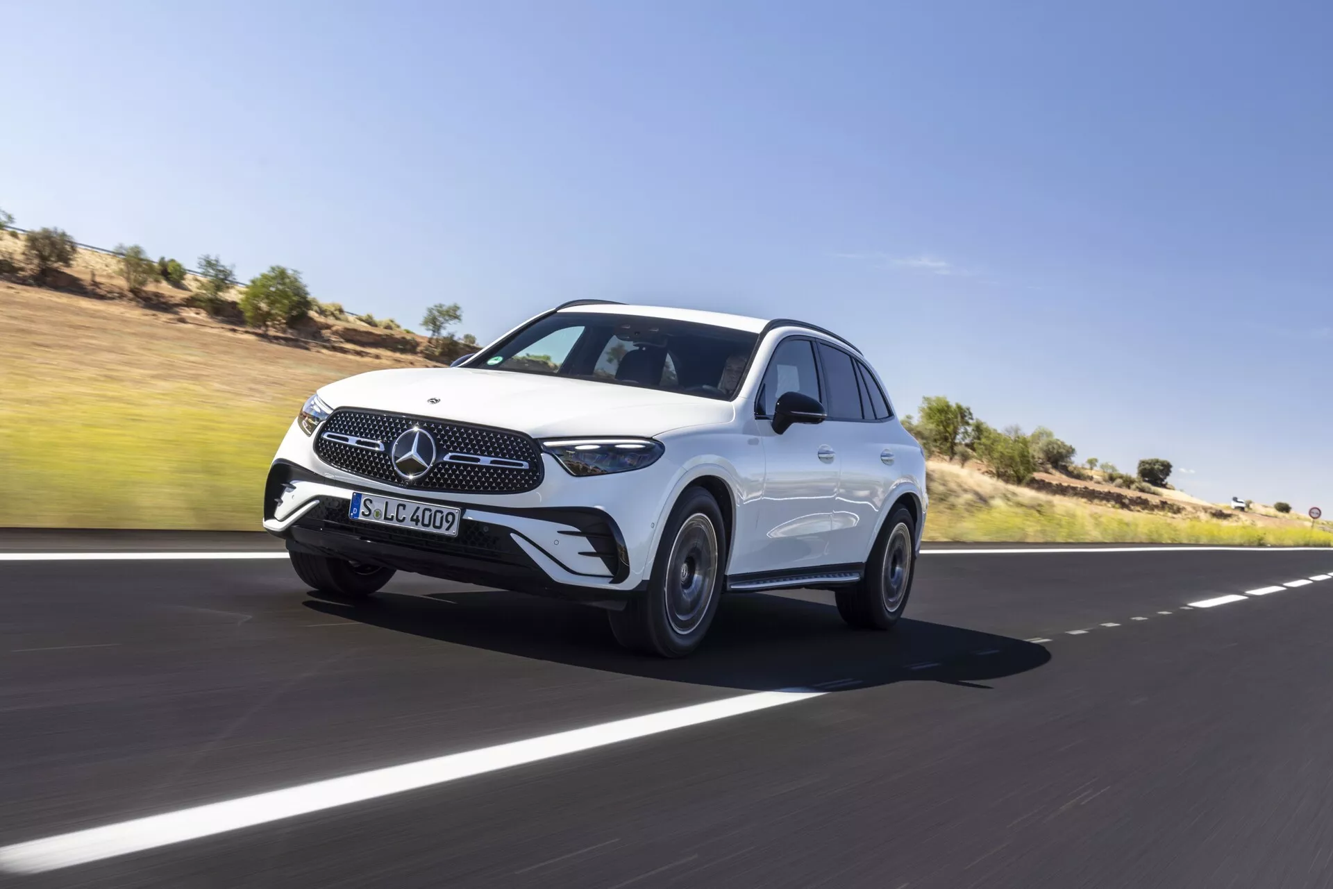 Dynamiczny kadr luksusowego SUV-a Mercedes GLC 2022 poruszającego się po gładkiej asfaltowej drodze, ukazujący jego przedni i boczny profil. Cechy obejmują charakterystyczną atrapę chłodnicy z trójramienną gwiazdą, eleganckie reflektory LED, czujniki na przednim zderzaku sugerujące zaawansowane systemy wspomagania kierowcy, rzeźbione linie maski, felgi aluminiowe, aerodynamiczne lusterka boczne z wbudowanymi kierunkowskazami i płynną linię dachu na sportową sylwetkę. Dominująca sylwetka pojazdu i niewielkie rozmycie wokół kół podkreślają jego ruch. Tło to czyste niebo i pustynny krajobraz.
