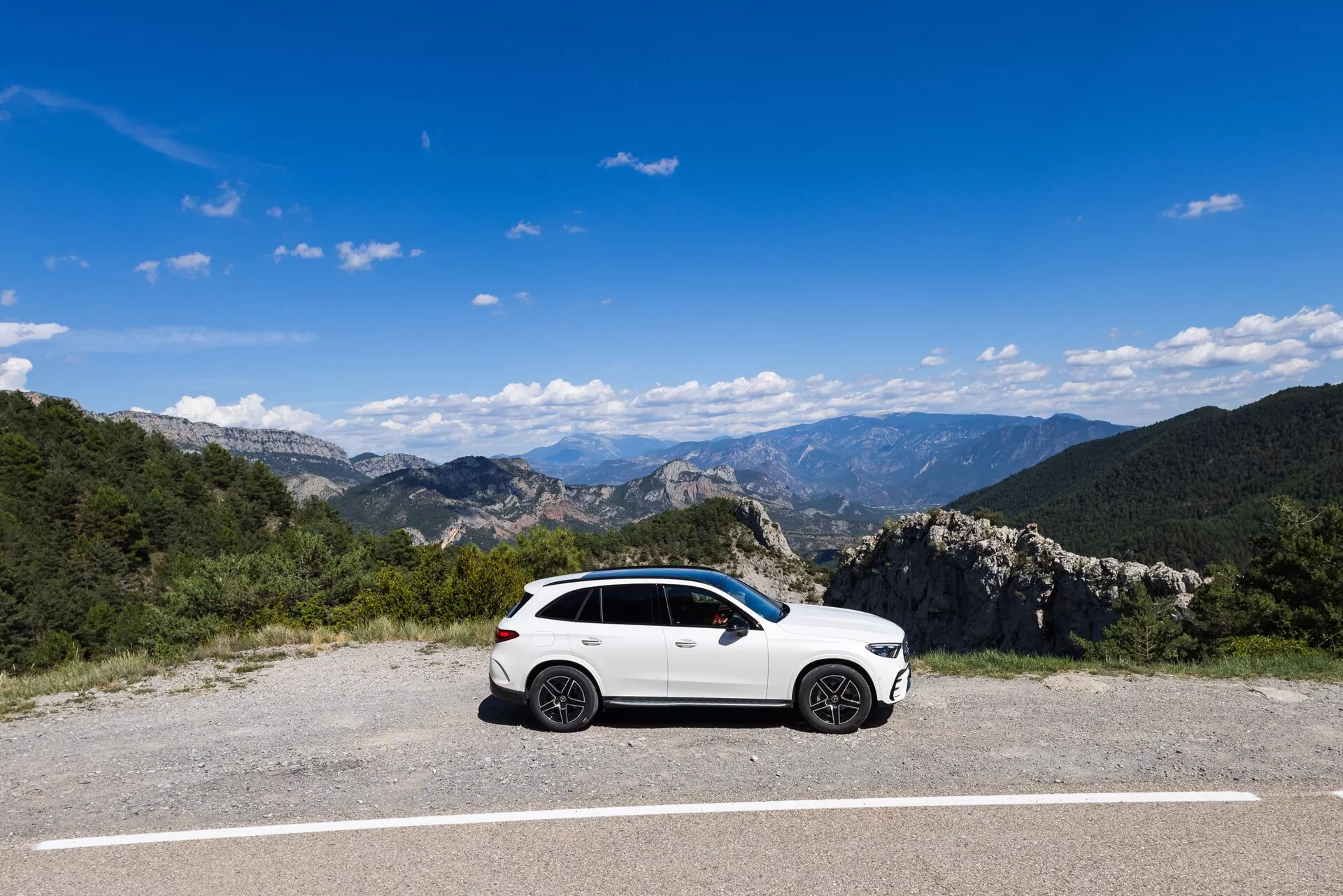 Białe, sportowe SUV Mercedes GLC 2022 zaparkowane na płaskiej, wybrukowanej powierzchni z naturalnym, górskim krajobrazem w tle, pod czystym, niebieskim niebem. Widok z profilu bocznego ukazuje stronę kierowcy pojazdu, podkreślając elementy projektowe takie jak alufelgi, lusterka boczne, wykończenia okien i linie nadwozia. Oświetlenie i klarowność umożliwiają widoczność subtelnych konturów na drzwiach bocznych.