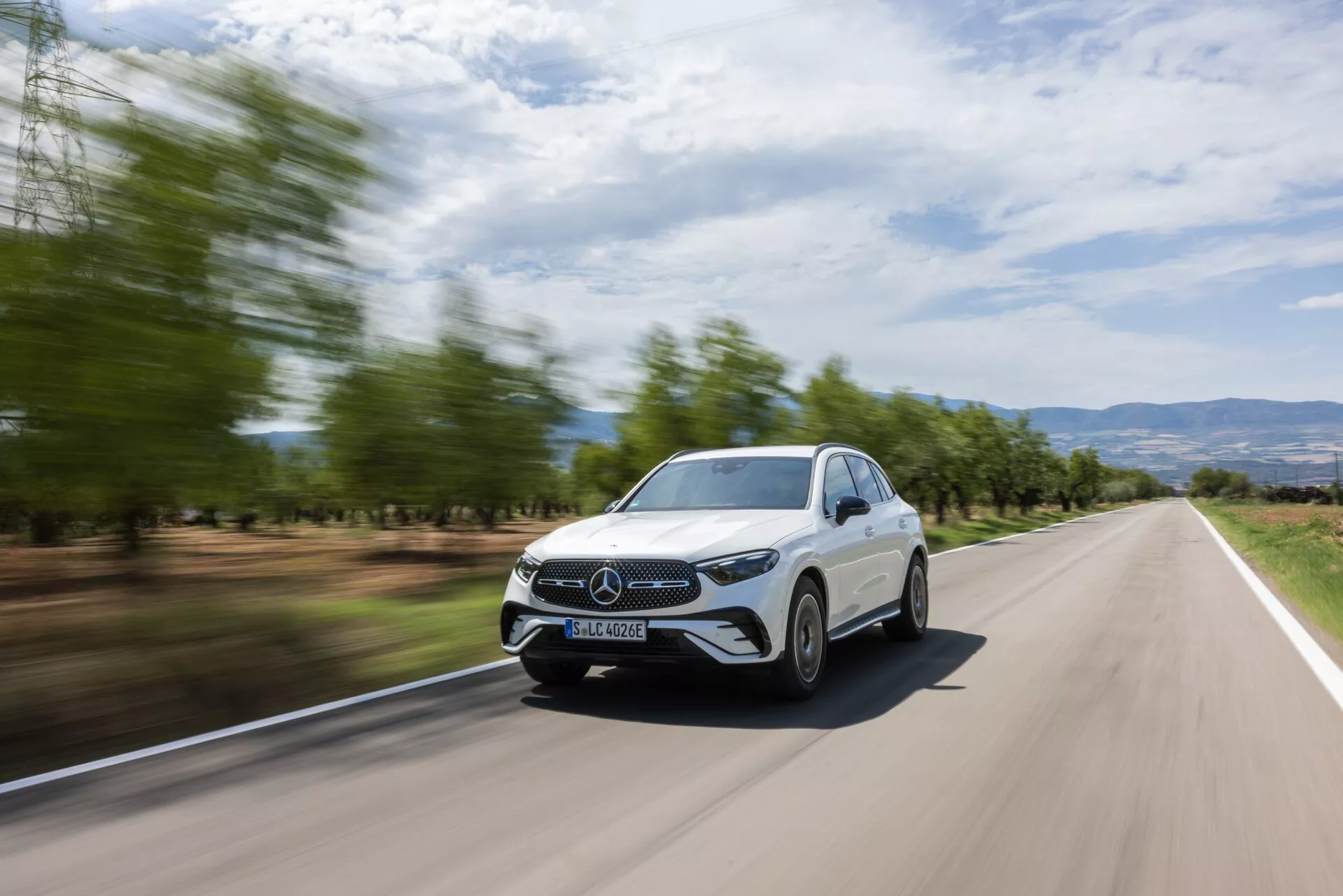 Widok z przodu pod kątem trzech czwartych na białego Mercedesa GLC 2022 w ruchu na dwupasmowej drodze, ukazujący jego przedni grill, reflektory, część maski, przednią szybę i stronę kierowcy. Tło jest rozmyte przez ruch, co podkreśla prędkość samochodu. Elegancki design, zamknięty grill, energooszczędne reflektory LED i widoczne światła do jazdy dziennej, wraz z nowoczesnym krajobrazem z rozmytymi drzewami i pagórkowatym terenem, prezentują dynamiczny i stylowy wygląd, idealny dla materiałów promocyjnych.